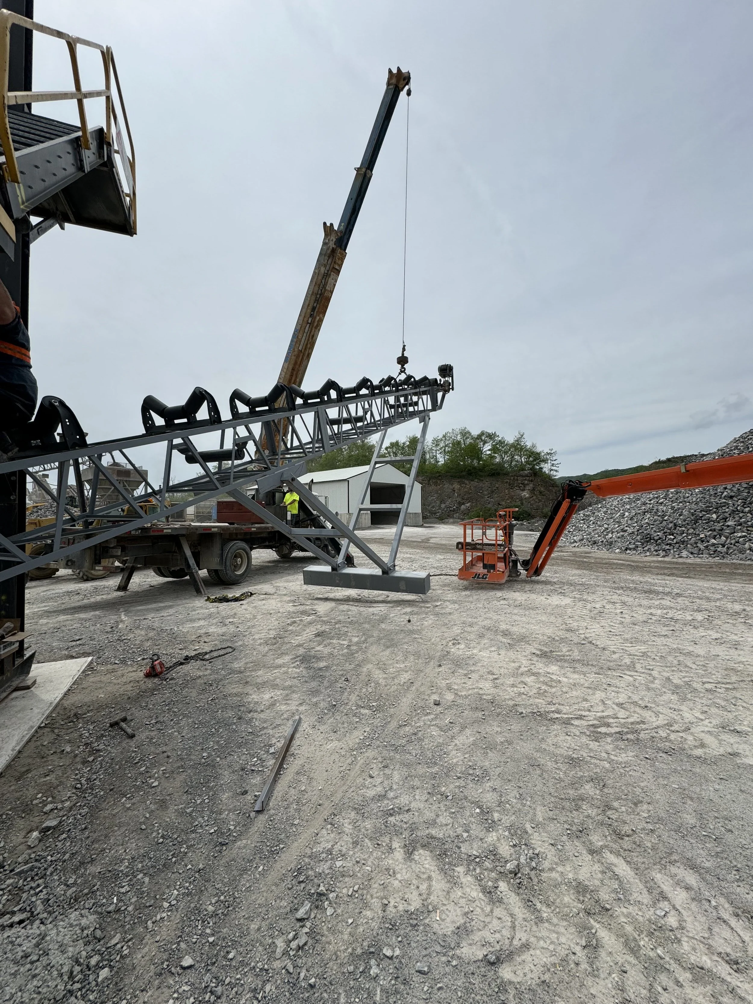 Construction site with heavy machinery, including a crane lifting a metal structure, and workers in safety vests. Gravel ground and a building in the background.