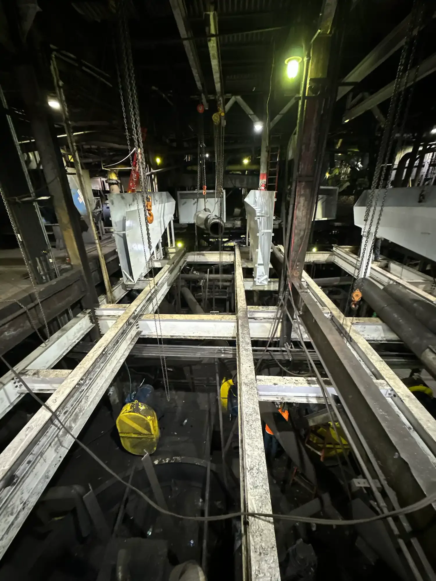 Inside a construction site or industrial facility with metal scaffolding, pipes, chains, and construction safety equipment, dimly lit with a greenish-yellow light.