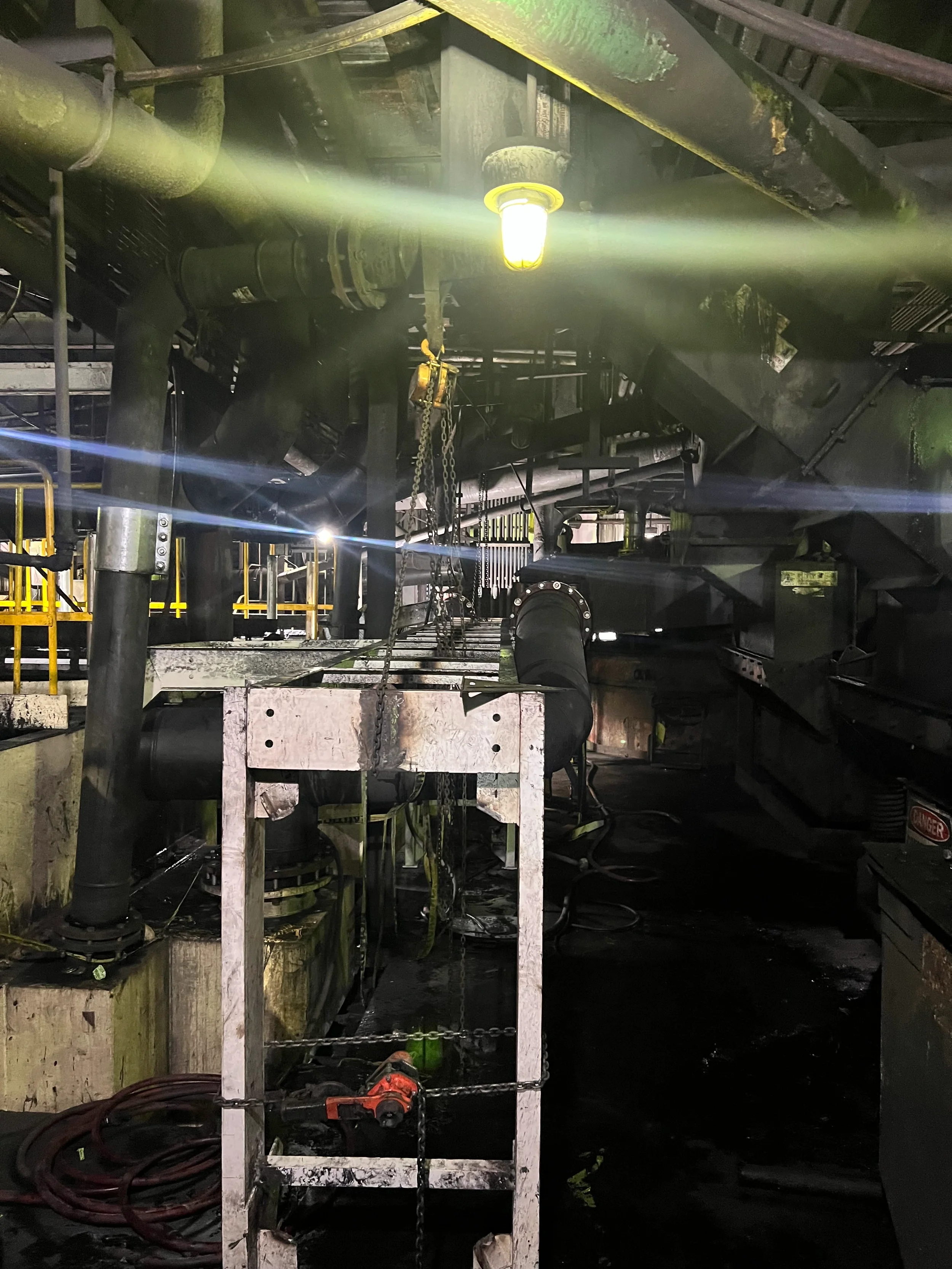 Industrial setting with large pipes, chains, and maintenance equipment, illuminated by overhead lighting, with safety barriers and a warning sign.