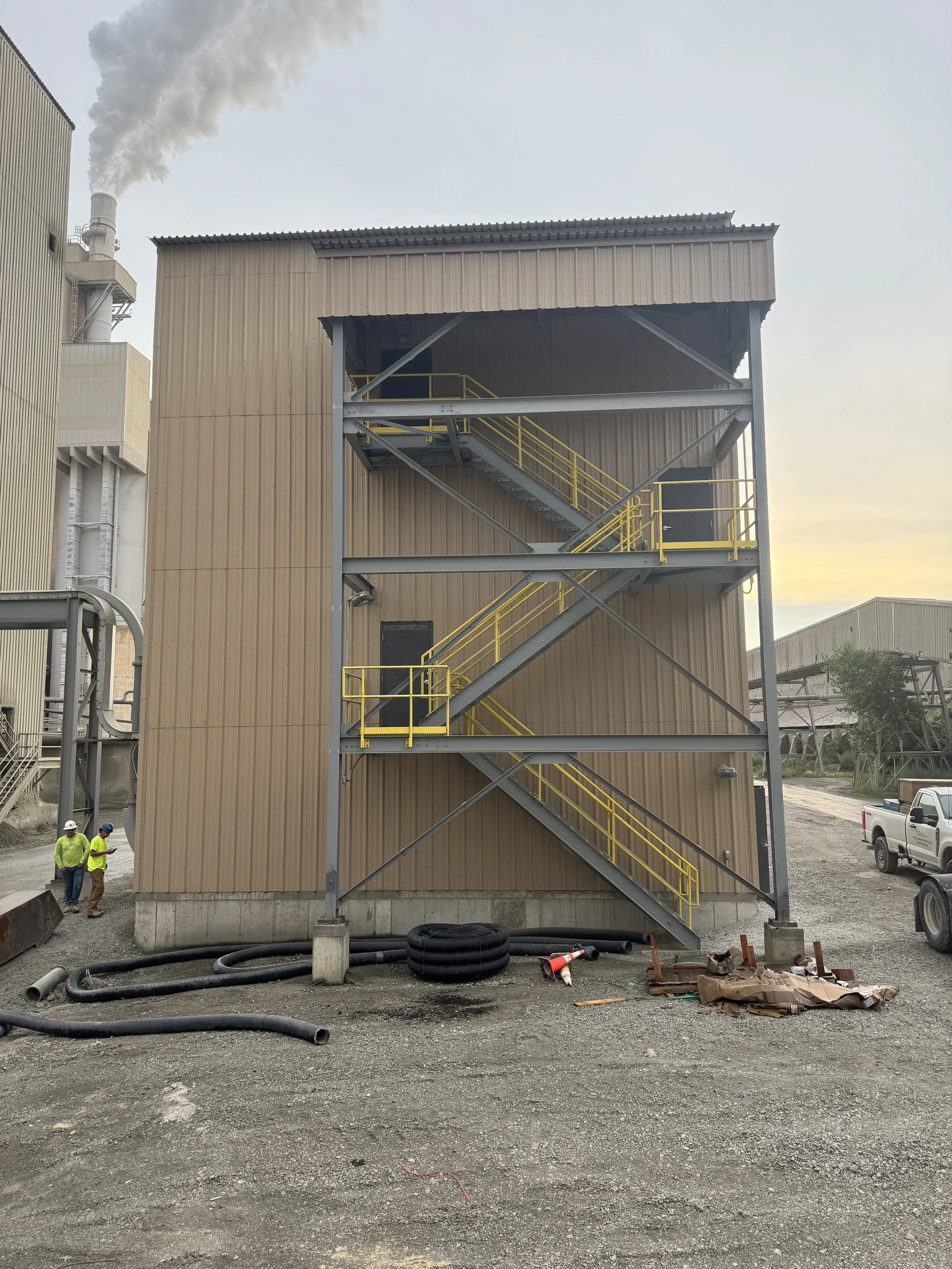 Exterior view of an industrial building with an external staircase and construction materials and pipes on the ground, with two workers wearing safety gear in the lower left corner.