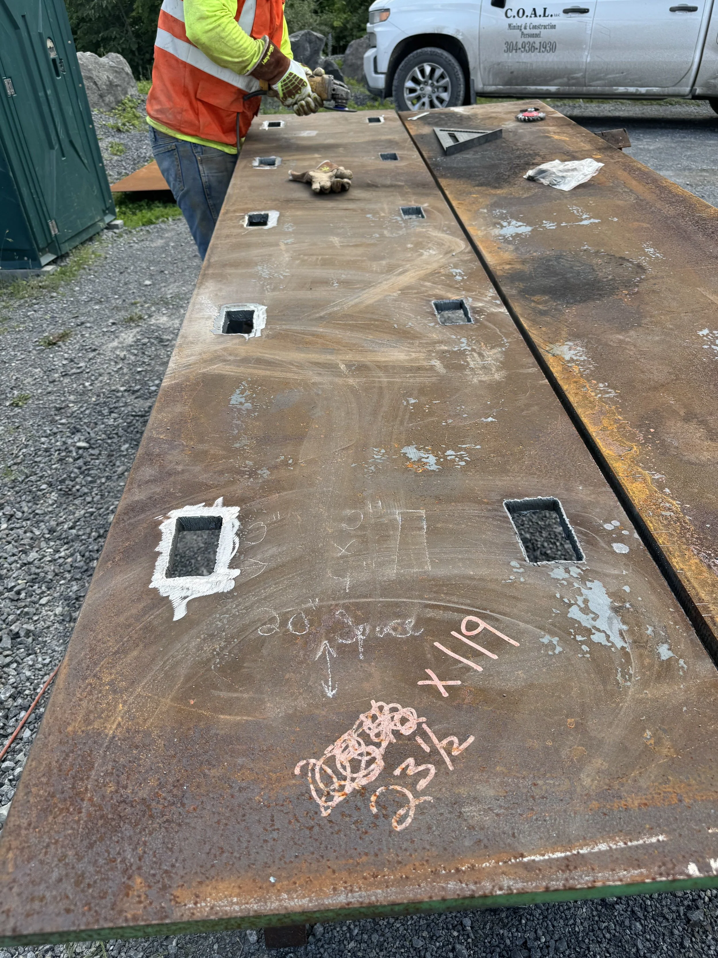 A work table with square holes and various markings and drawings, including a gear and wheels in chalk, outdoors with a person in a safety vest and gloves using a power tool.