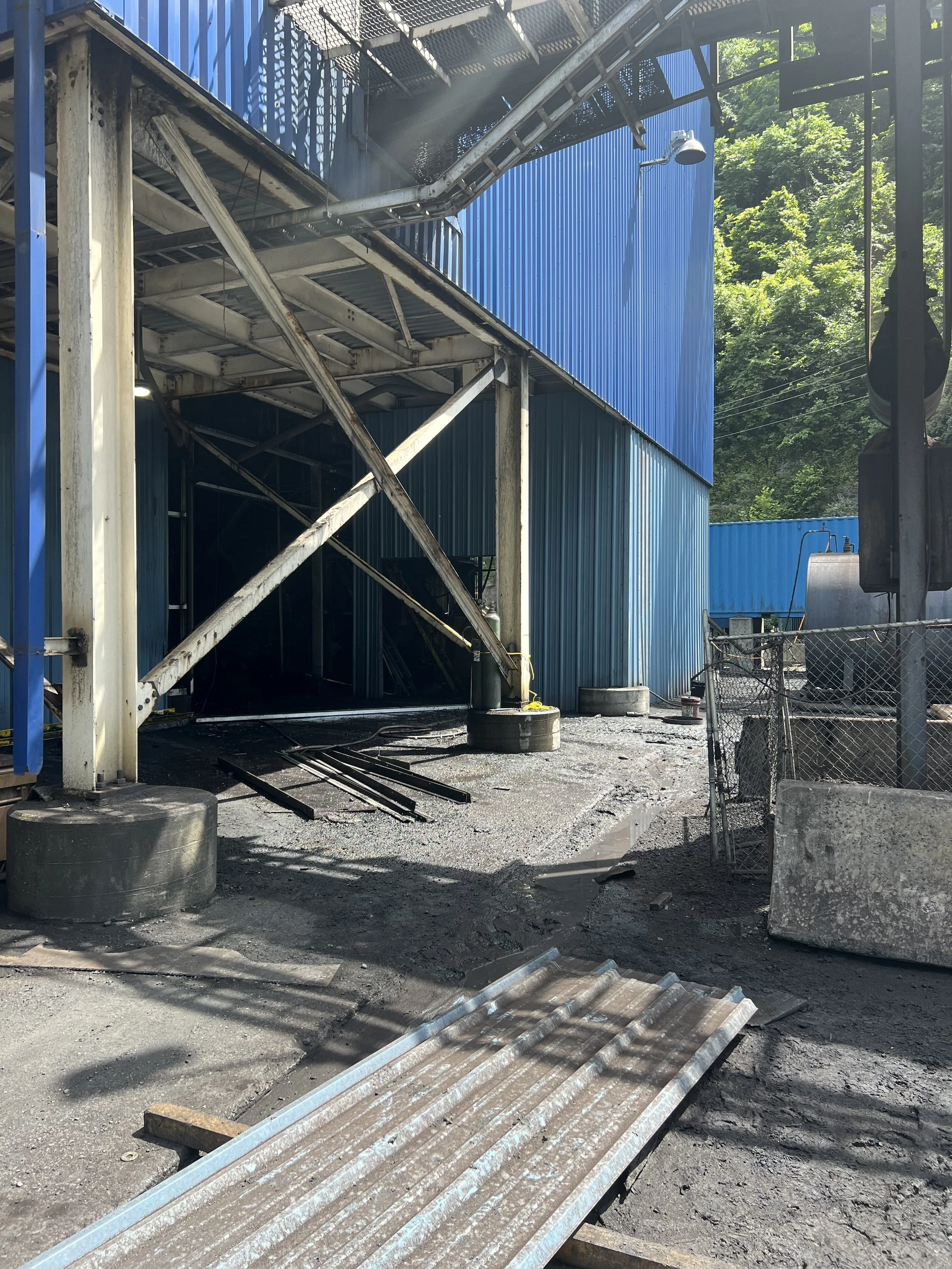 Construction site with a blue building, metal stairs, and scaffolding, surrounded by trees and a chain-link fence.