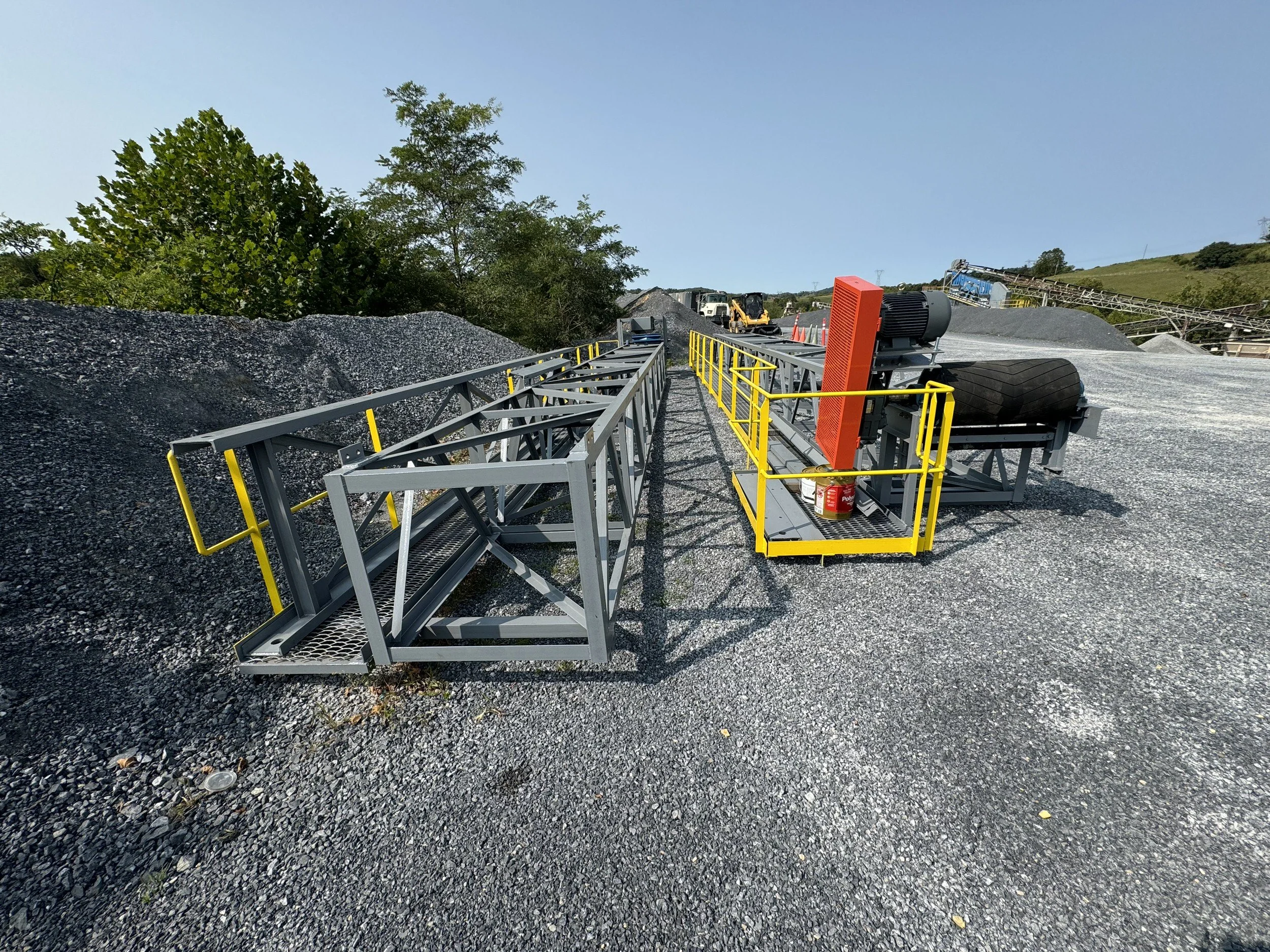 View of a construction or mining site with gravel piles, heavy machinery, a conveyor belt, and a blue sky with some trees in the background.