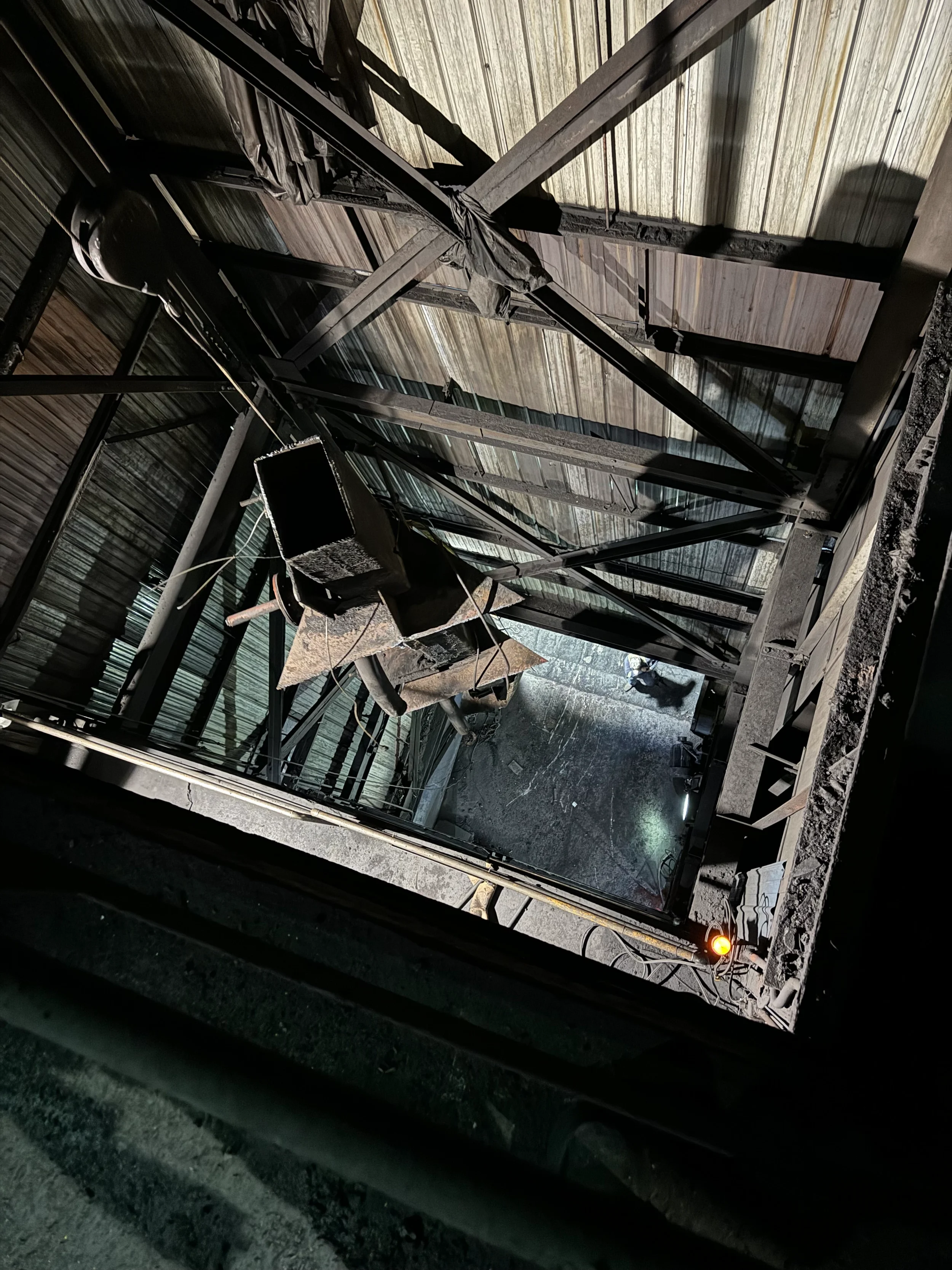 View from the inside of an elevator shaft showing the structural frame, wiring, and damaged or construction materials, with a person visible at the bottom.