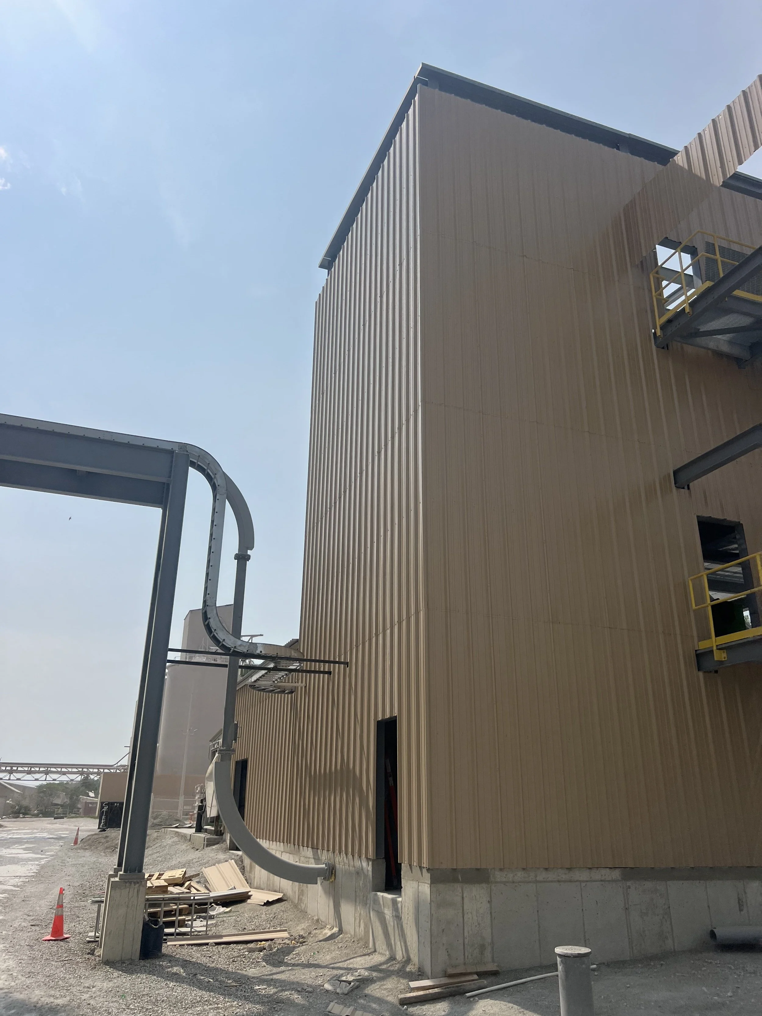 Construction site with a large, beige, vertically ridged building exterior, metal stairs with yellow railings, and industrial piping under a clear blue sky.
