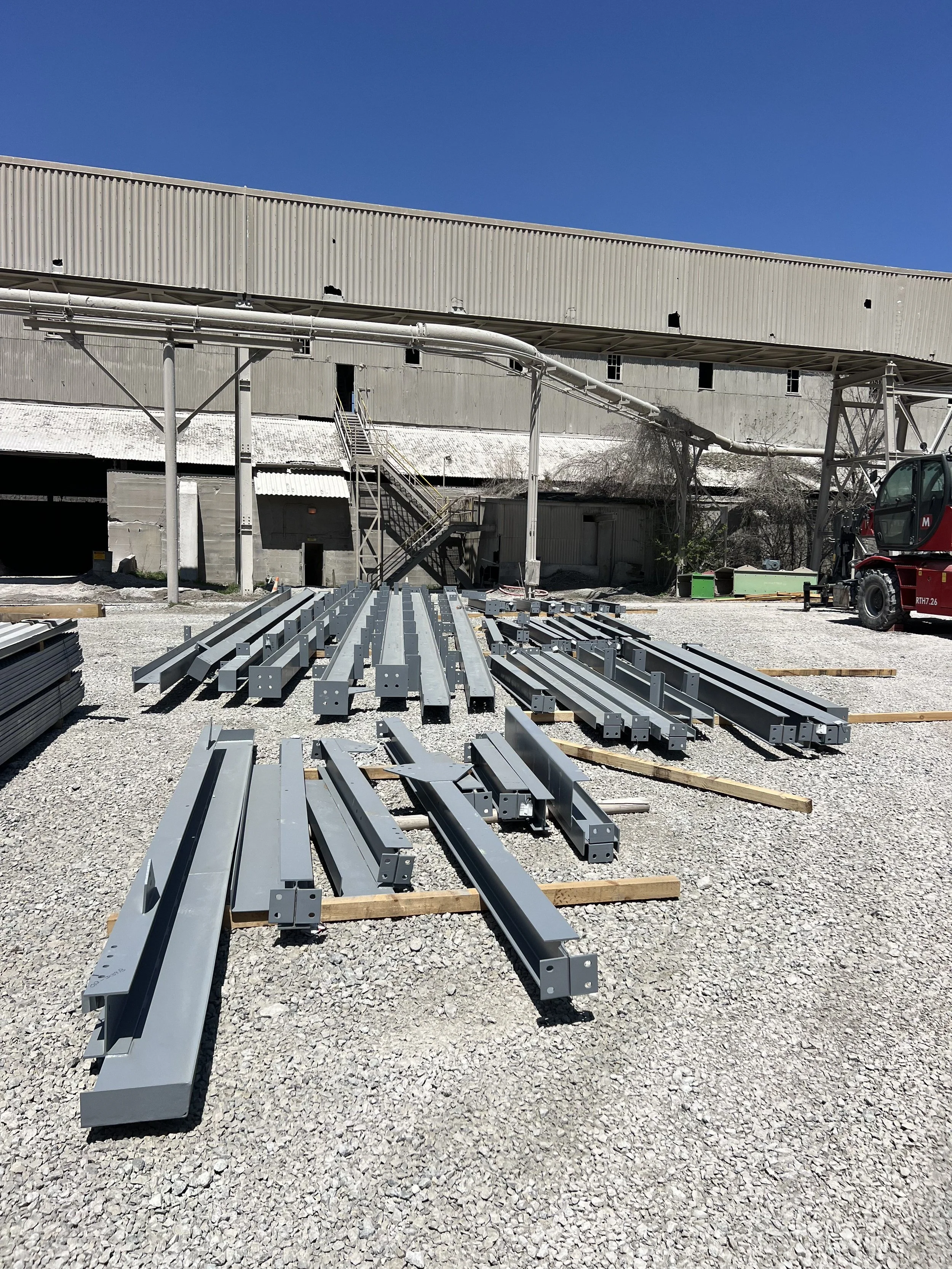 Construction site with metal beams on gravel ground, a large industrial building with holes in the exterior wall, and a small crane on the right.