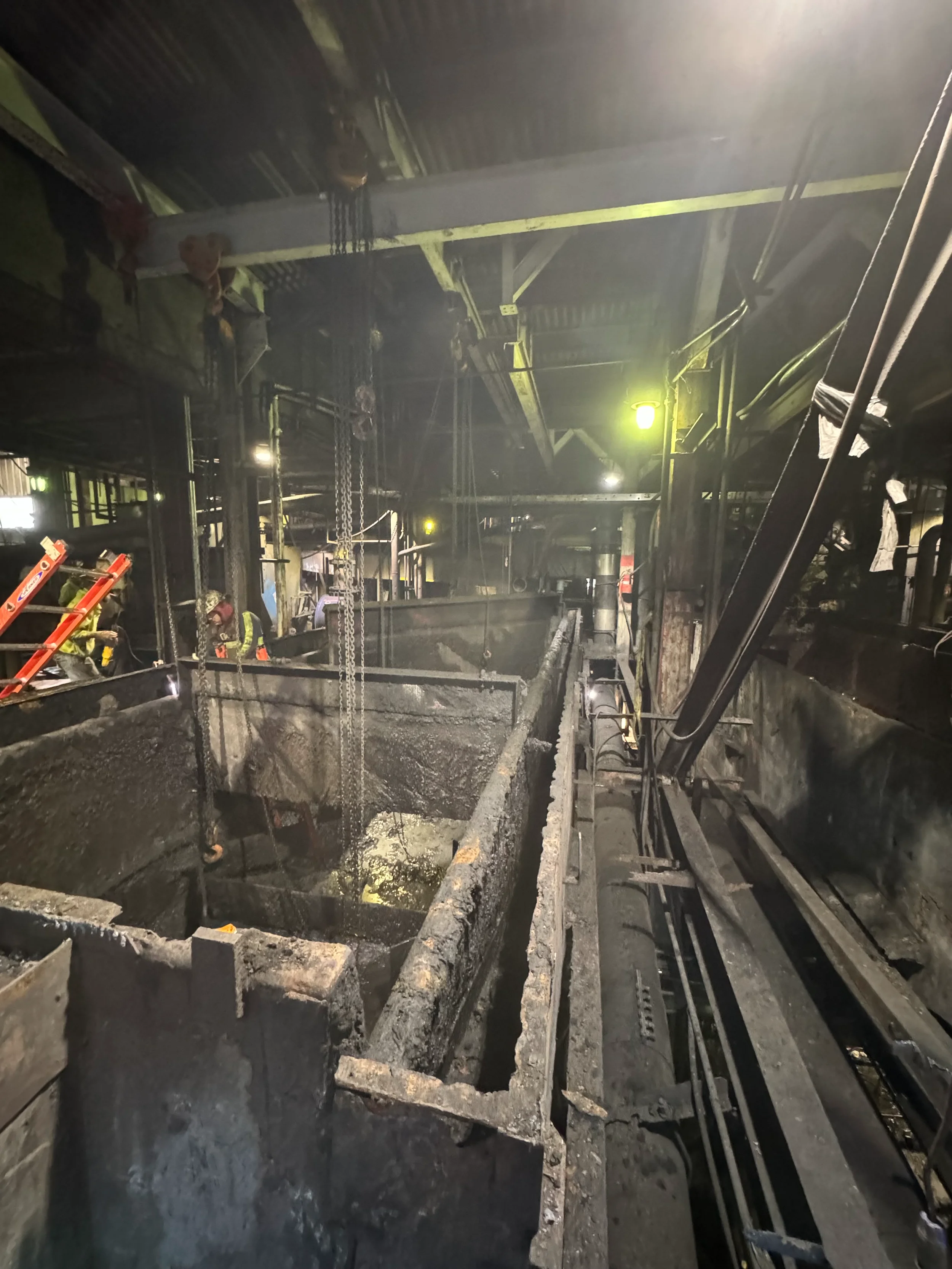 Construction workers wearing safety gear working inside a dark industrial facility with large machinery and structural beams, possibly involved in maintenance or repair tasks.