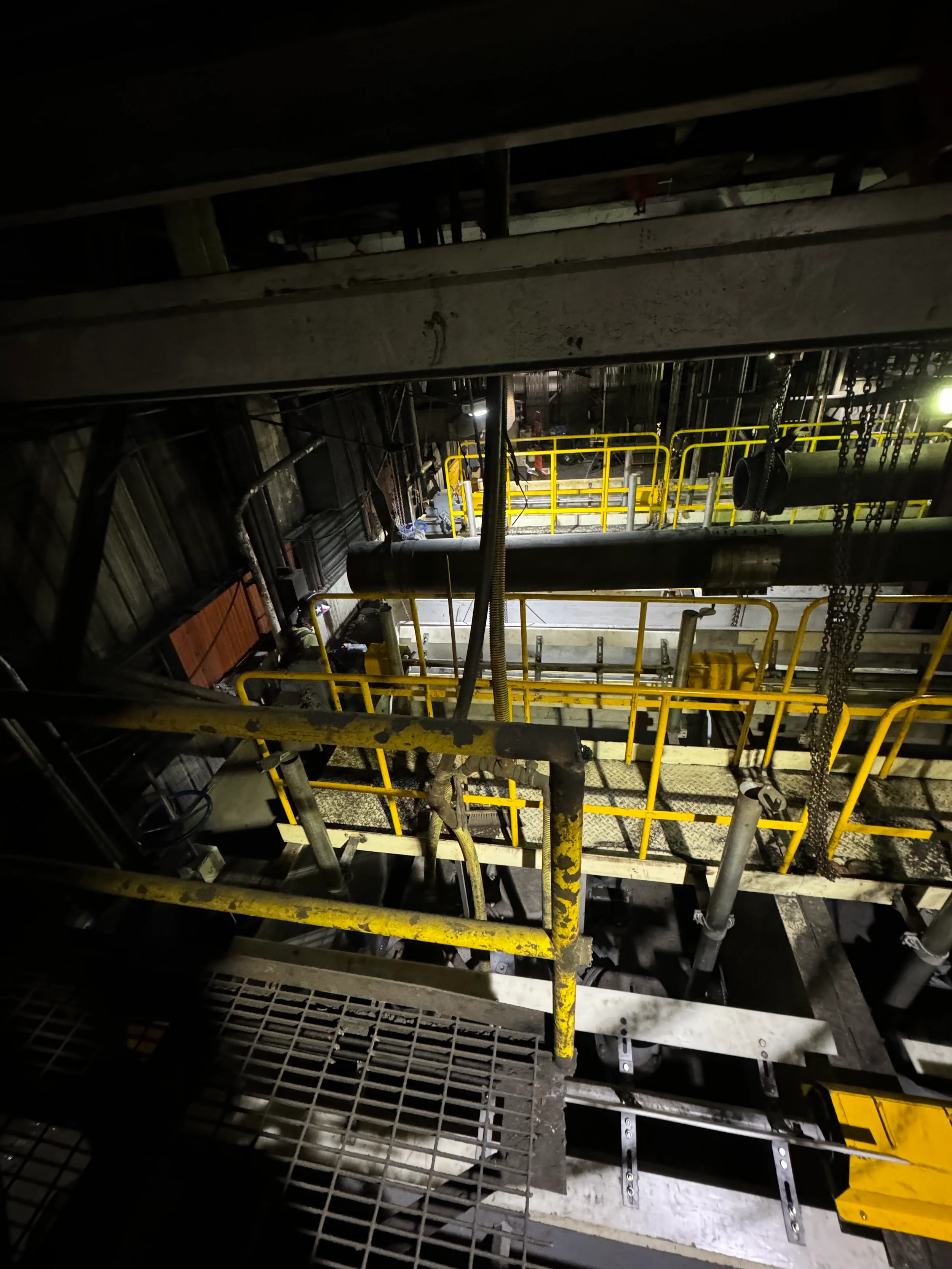 A view from below of an industrial or construction site with metal scaffolding, pipes, and yellow safety rails in a dark environment.