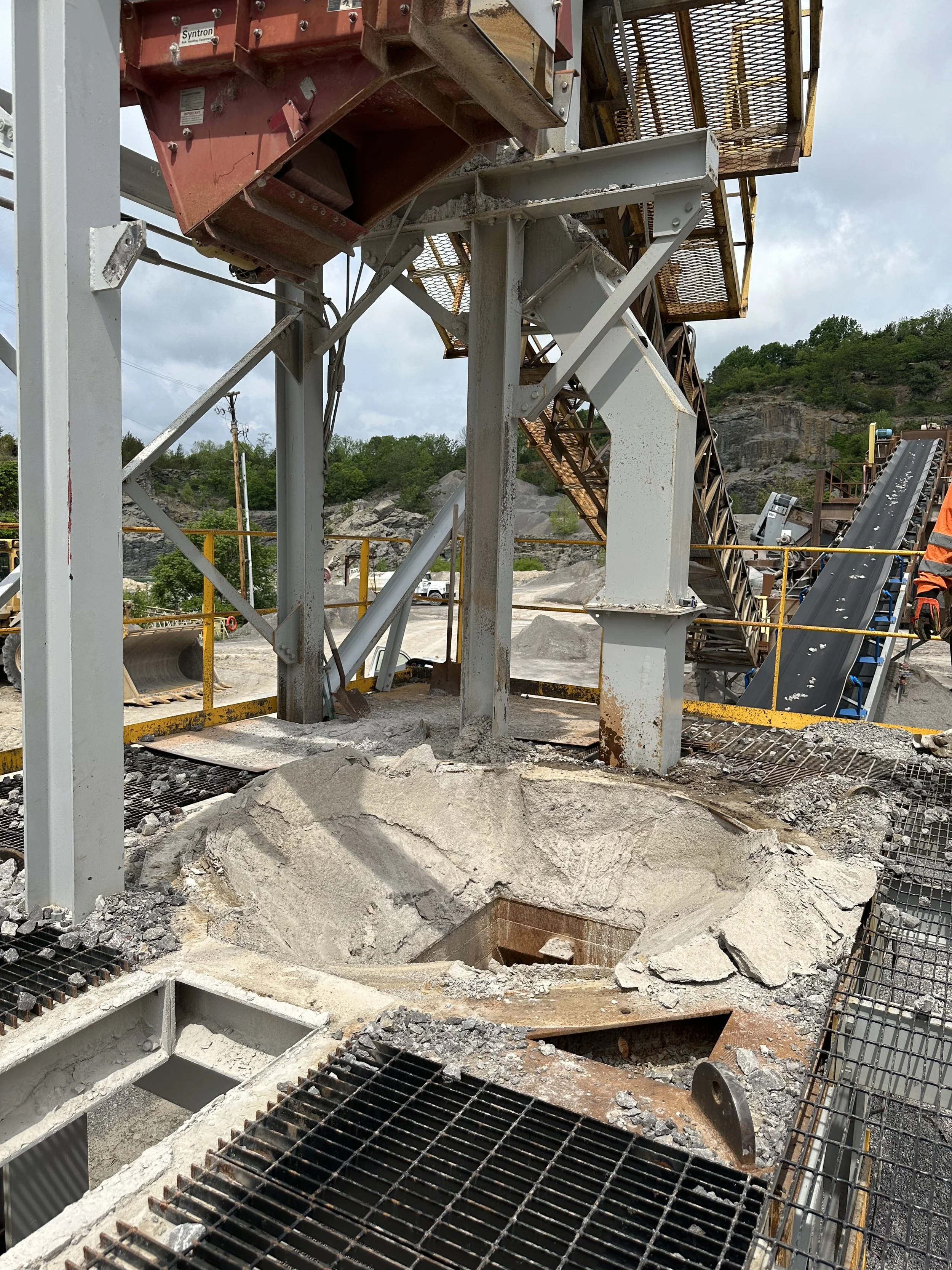 Construction site with metal framework, conveyor system, and cut rock surface. Overcast sky with hills in the background.