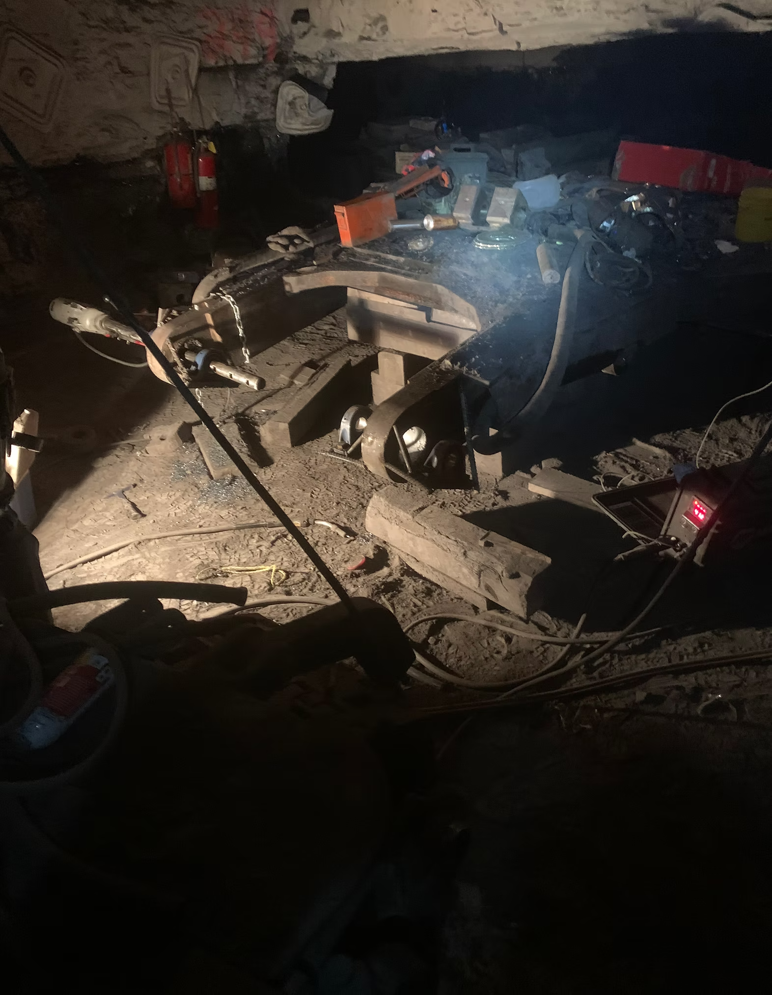 A cluttered workspace with tools, parts, and supplies scattered on a dirt floor, with a workbench in the background, illuminated by a bright light.