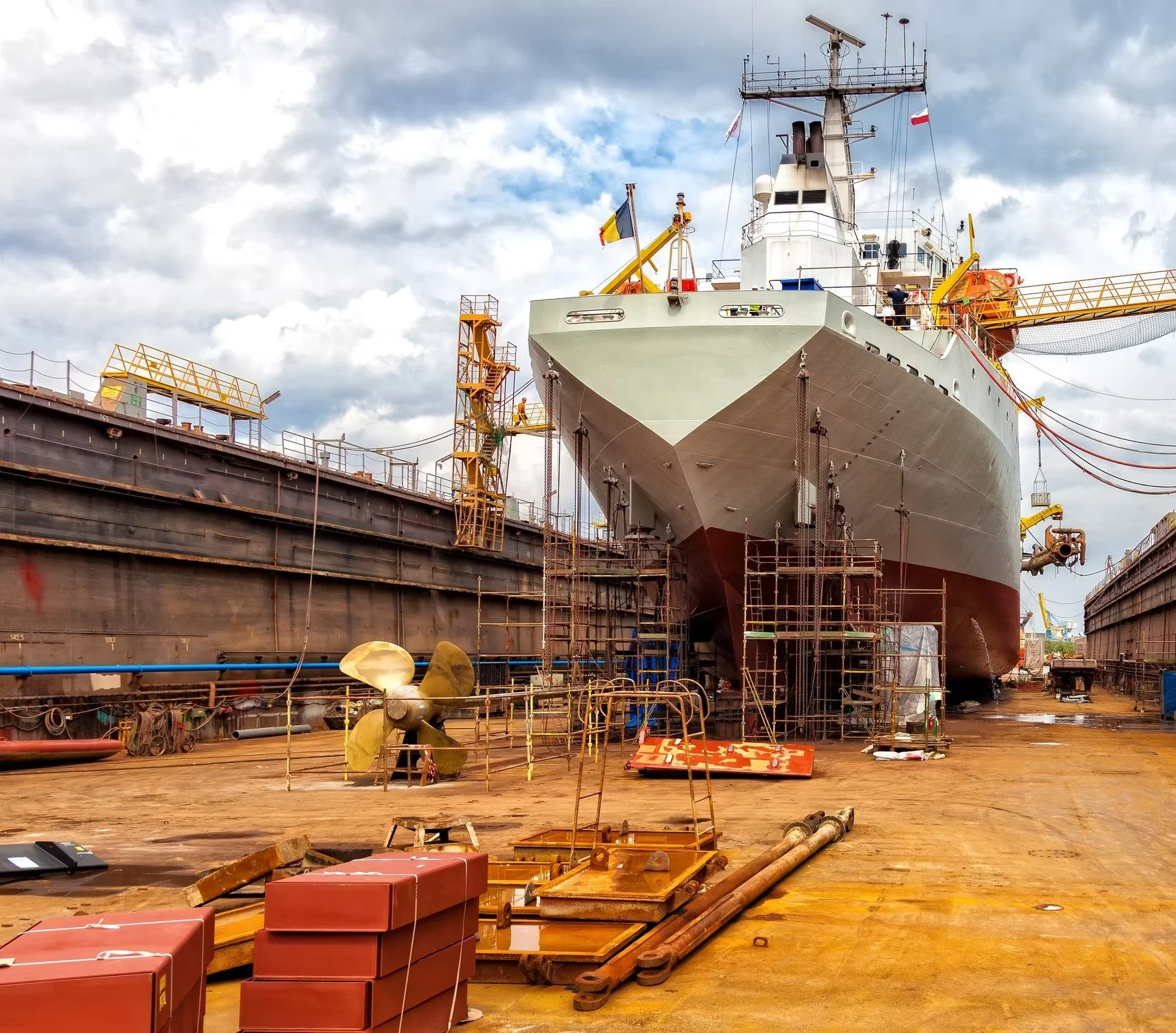 A large ship is in dry dock undergoing maintenance, surrounded by scaffolding and construction equipment, with a cloudy sky overhead.