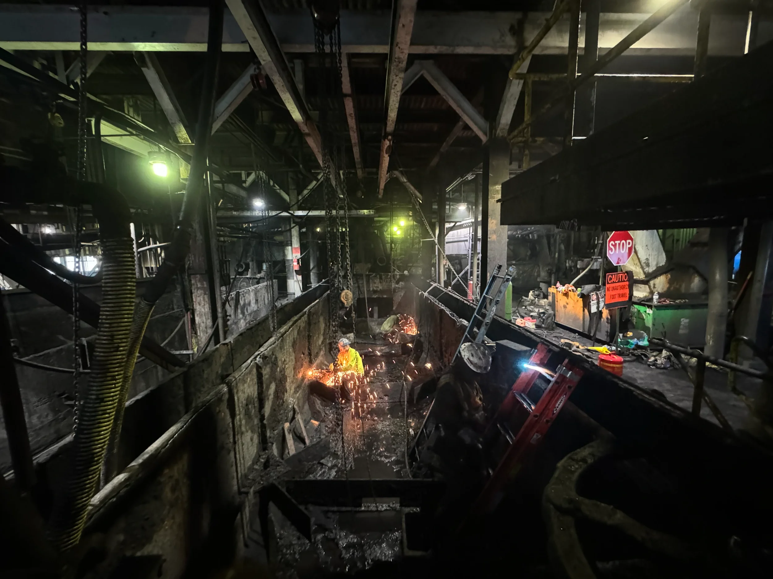 Workers in safety gear welding inside an industrial construction site at night, with scaffolding, chains, and safety signs visible.