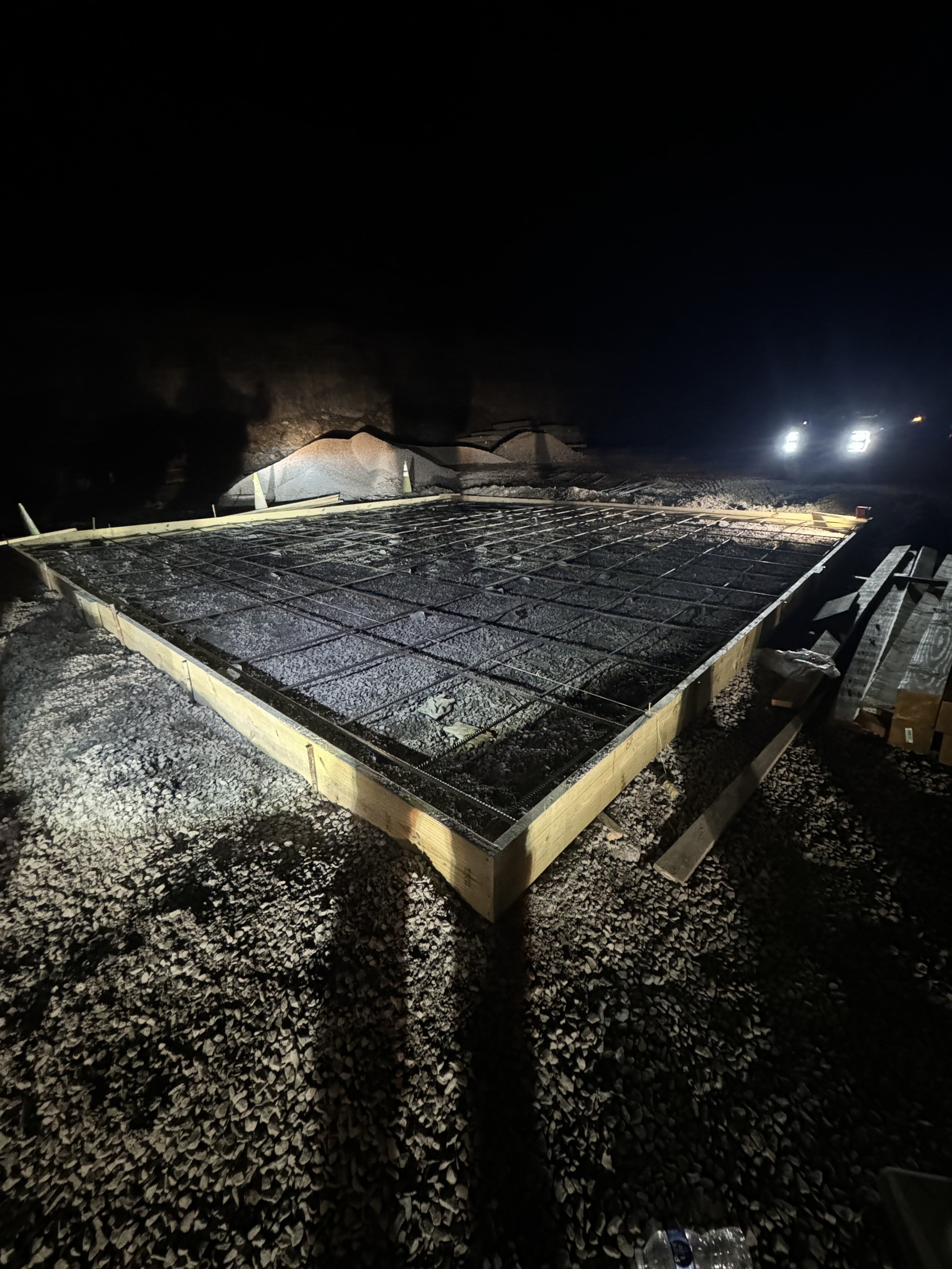 Construction site at night with a foundation frame and rebar, gravel on the ground, and bright headlights in the background.
