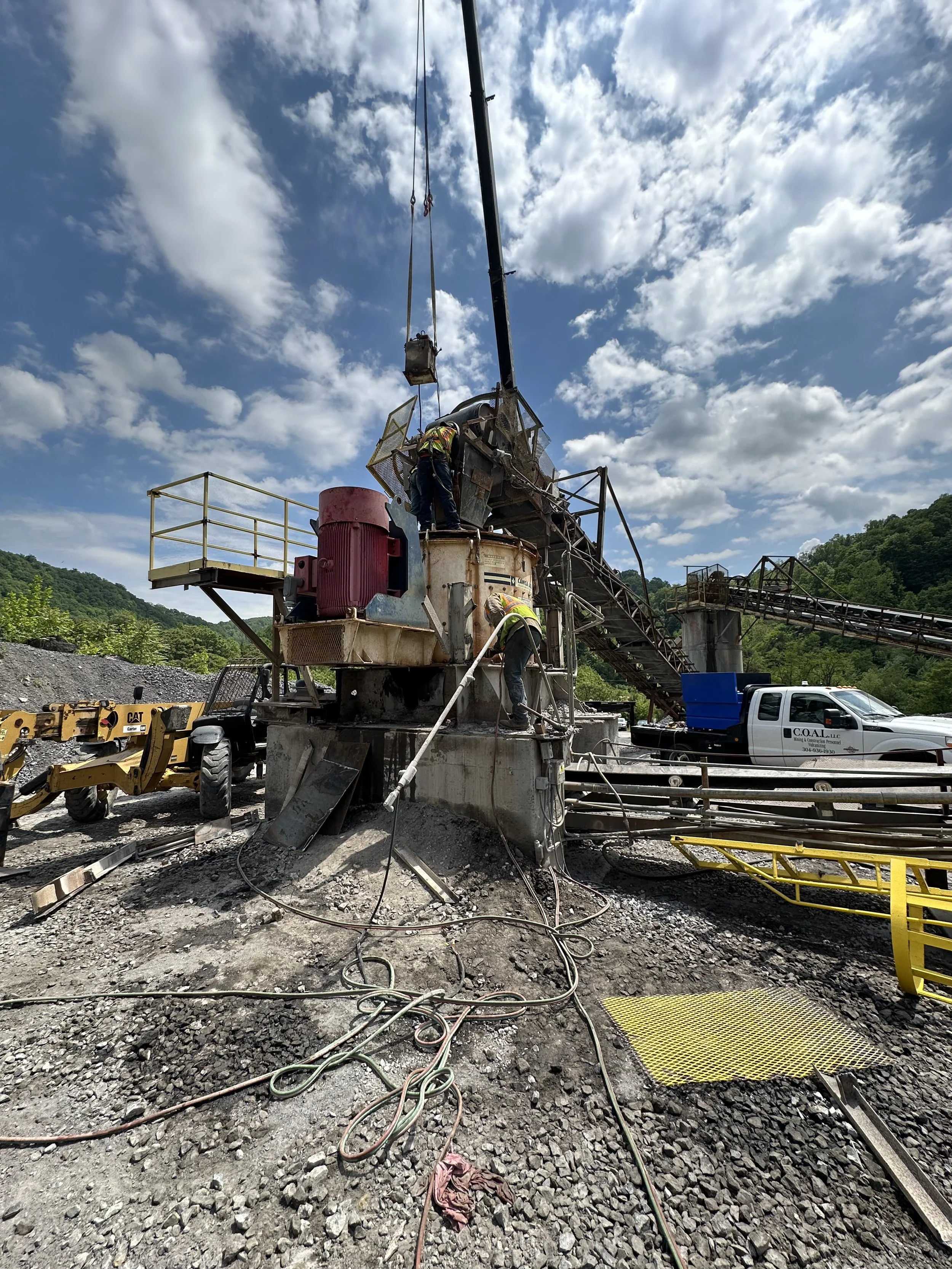 Workers operating a large industrial drilling machine outdoors under a blue sky with clouds, surrounded by rocky ground and construction equipment.