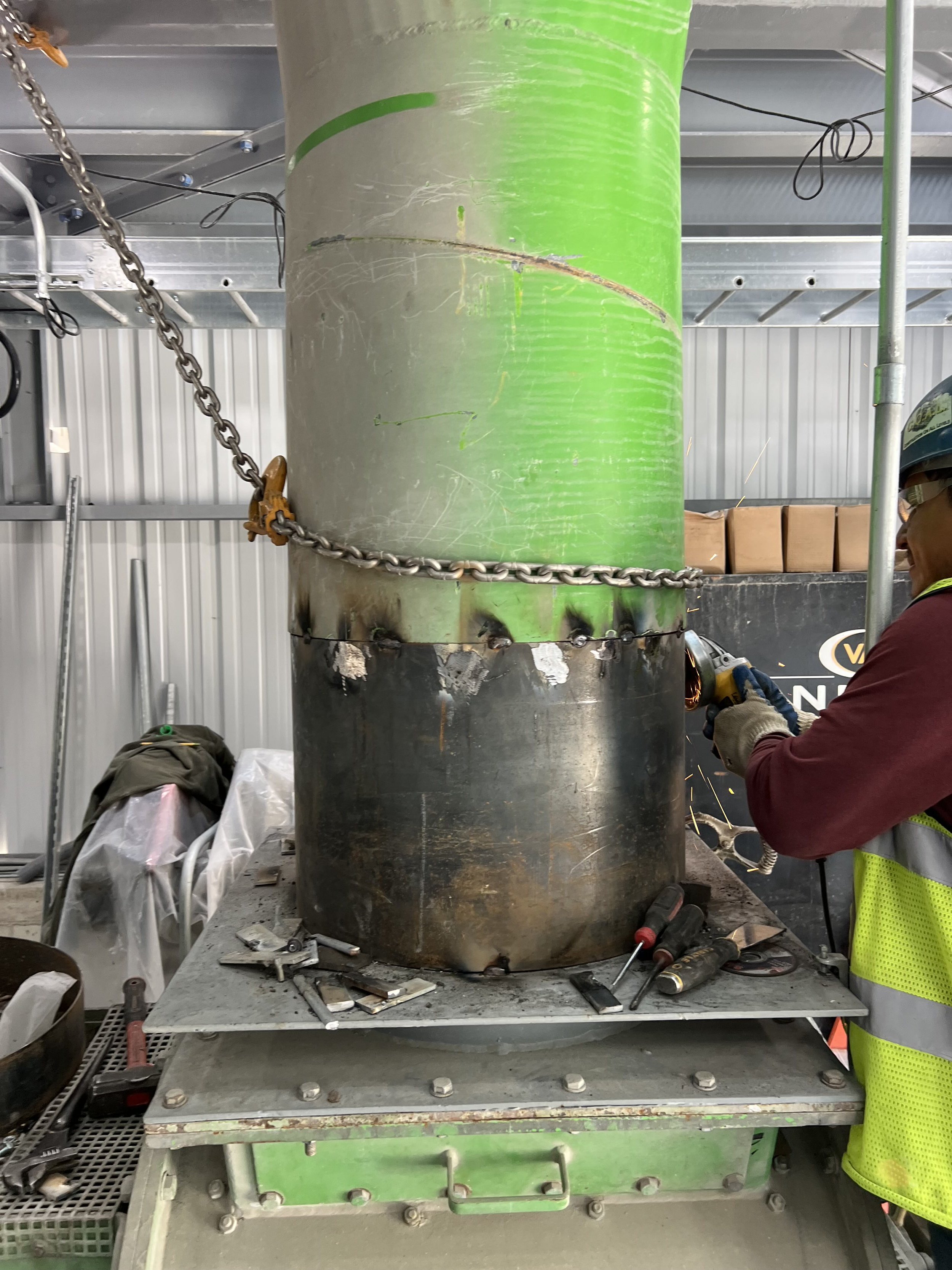 A worker welding a large green pipe with sparks flying, surrounded by tools and equipment in an industrial setting.