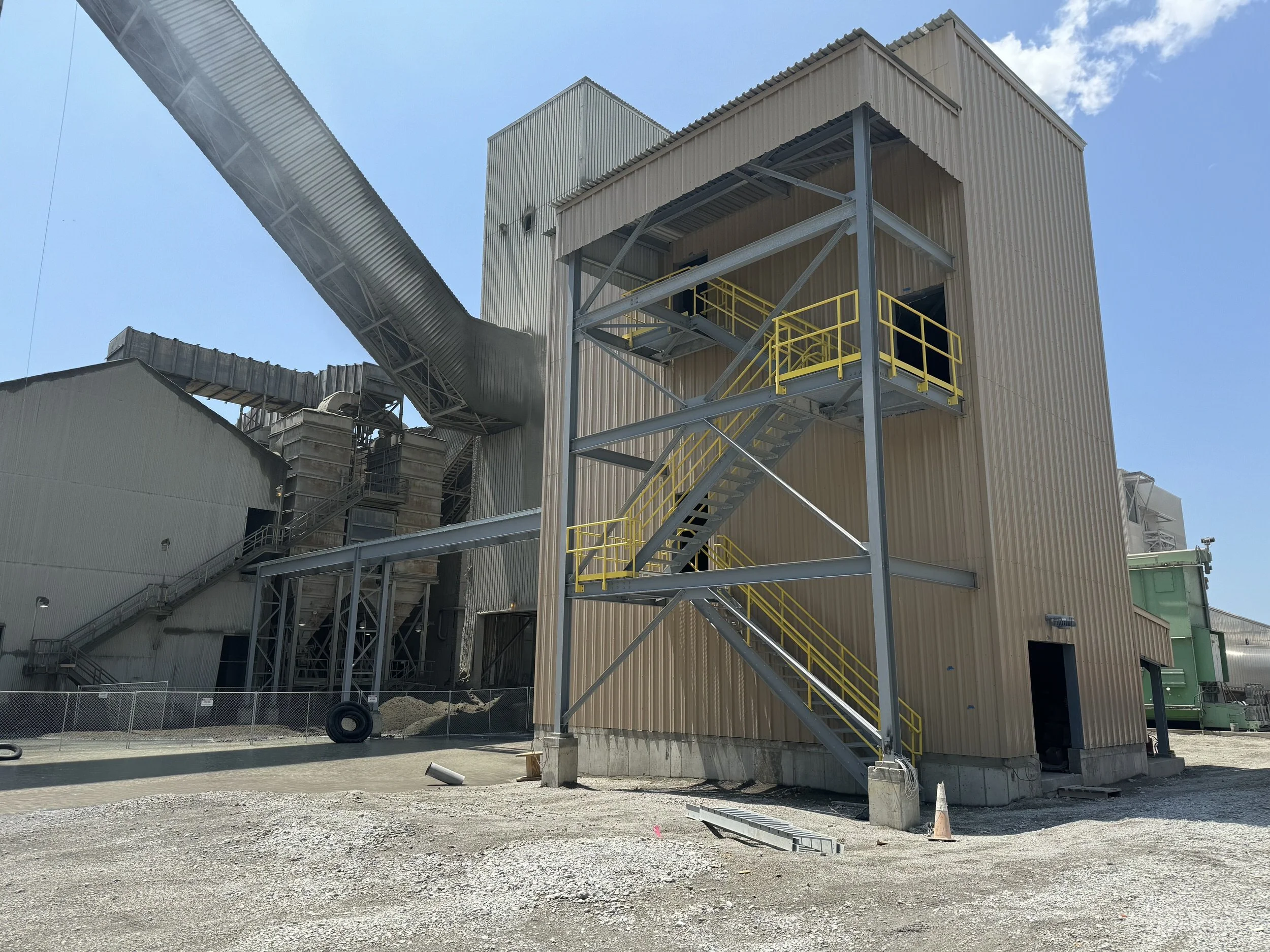 Industrial building under construction with metal stairs, yellow safety railings, large ductwork, and gravel ground against a blue sky with some clouds.