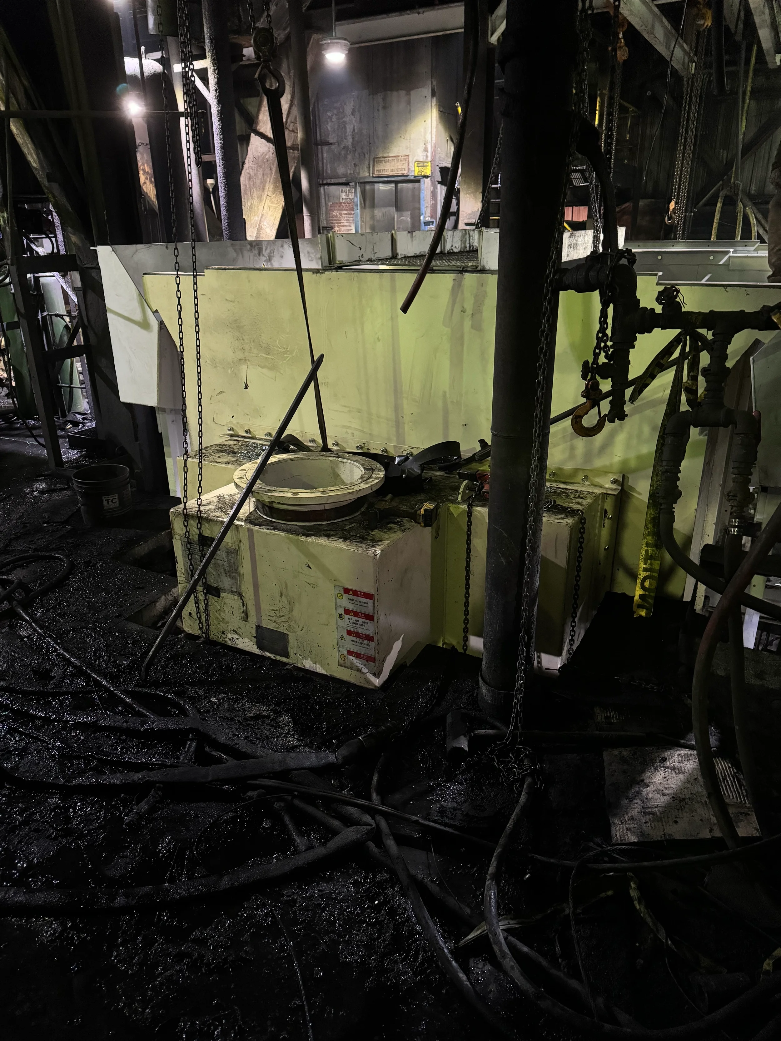 Image of industrial machinery in a dark, damaged environment with melted blackened surfaces, welded metal components, and chains hanging from the structure.