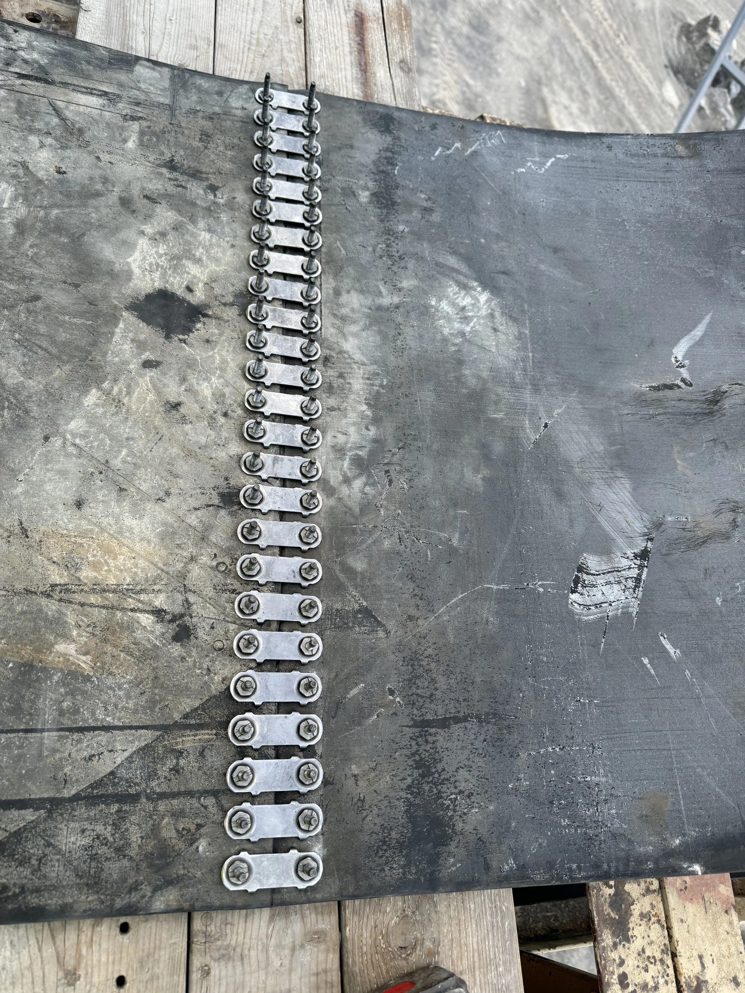 Metal mechanical part with bolts and links on a black surface, possibly part of a chain or a mechanical assembly. Wooden planks partially visible beneath.