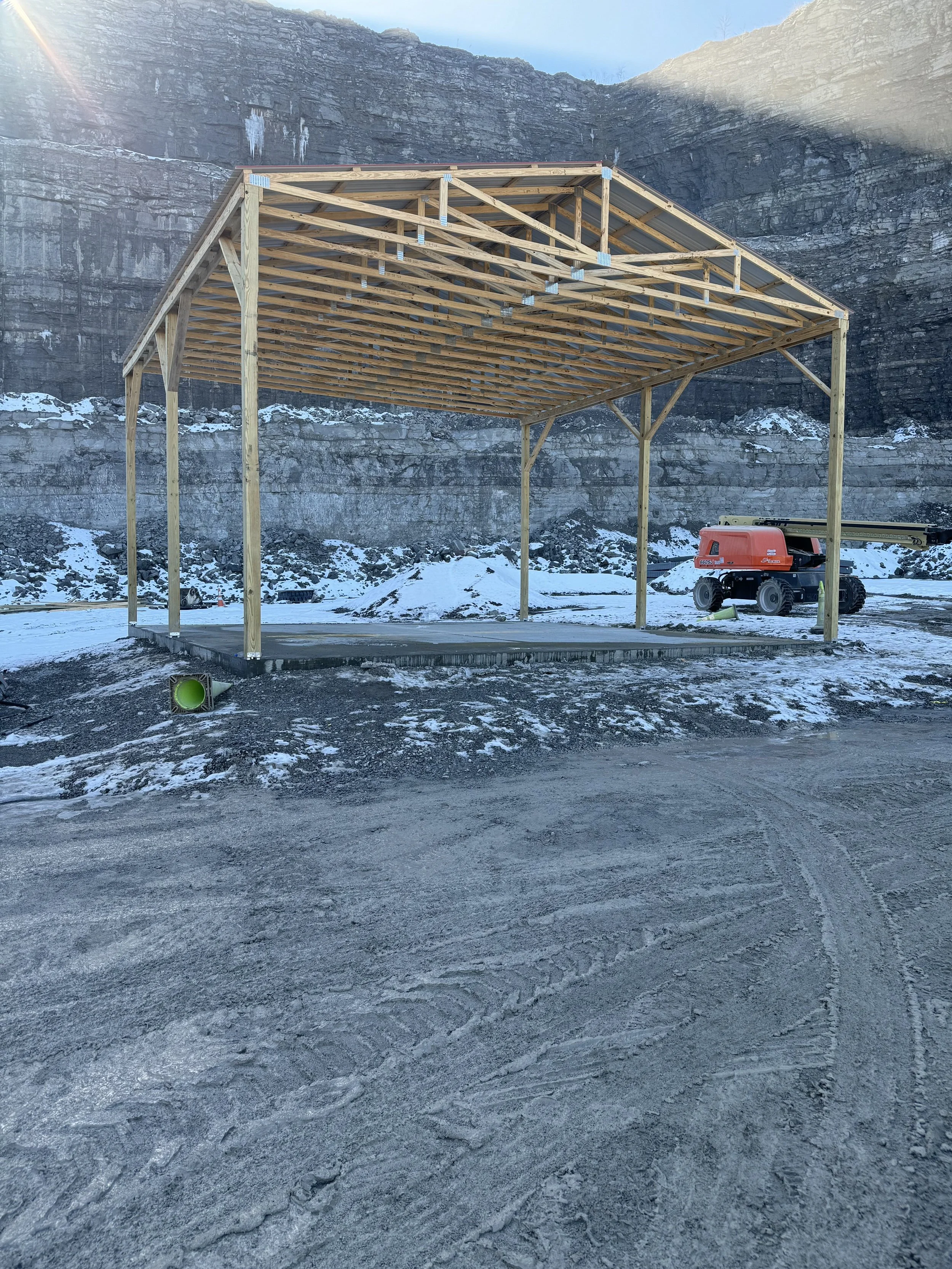 Construction site with a partially built wooden structure, a cherry picker, and snow on the ground, with a rocky cliff in the background.