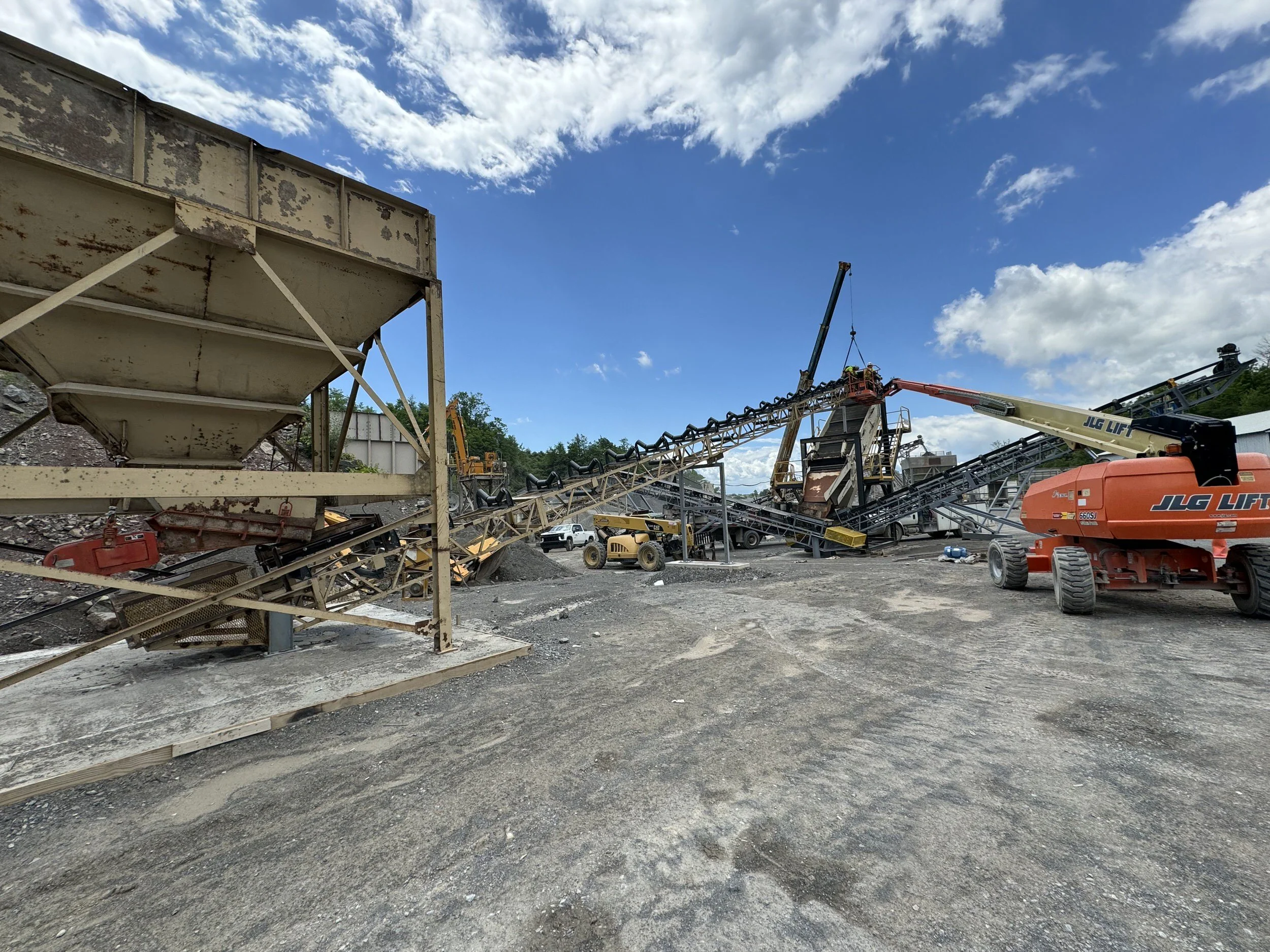 Construction site with machinery including a crane lifting an object, conveyor belts, and a JLG lift, with a cloudy blue sky overhead.