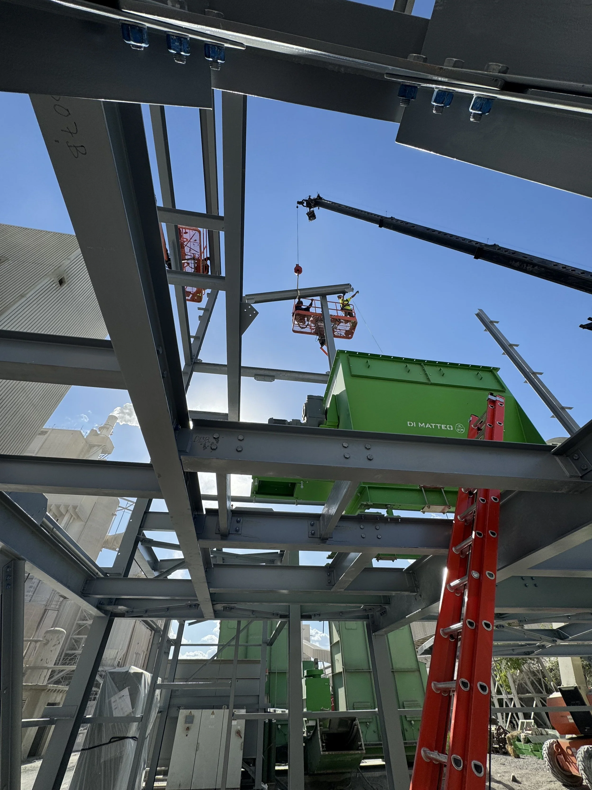 Construction site with a crane lifting a metal beam, workers on a cherry picker, red ladders, and metal framework against a blue sky.