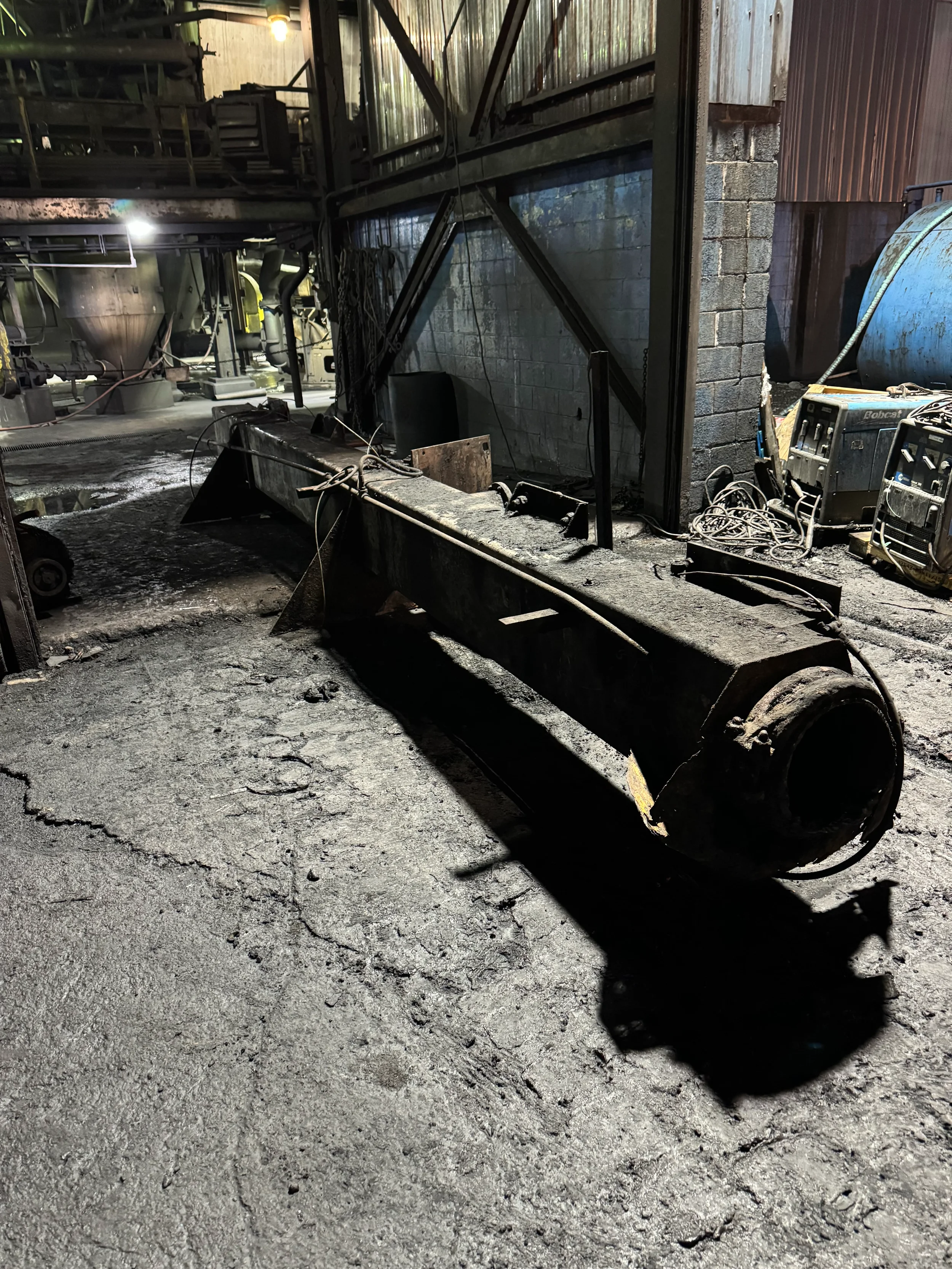 An industrial or factory setting with charred or burnt machinery, cables, and equipment on a dirty floor, indicating recent fire damage.