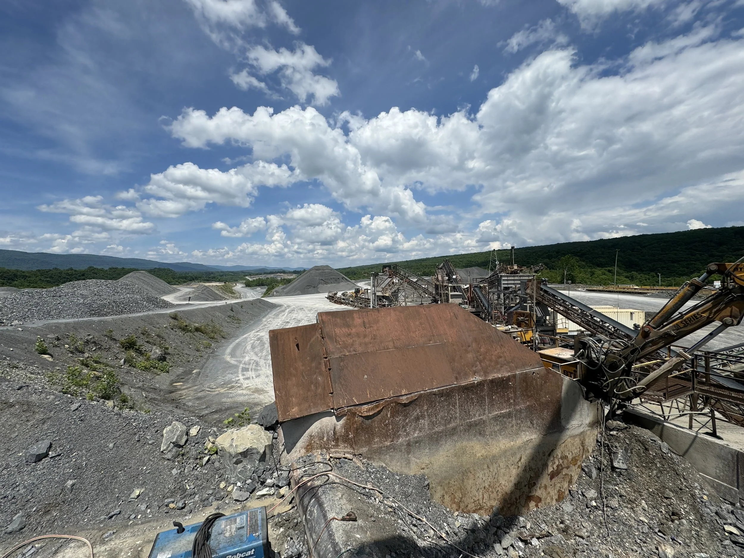 A large industrial site with gravel piles, machinery, and conveyor belts under a partly cloudy sky, surrounded by green hills in the distance.