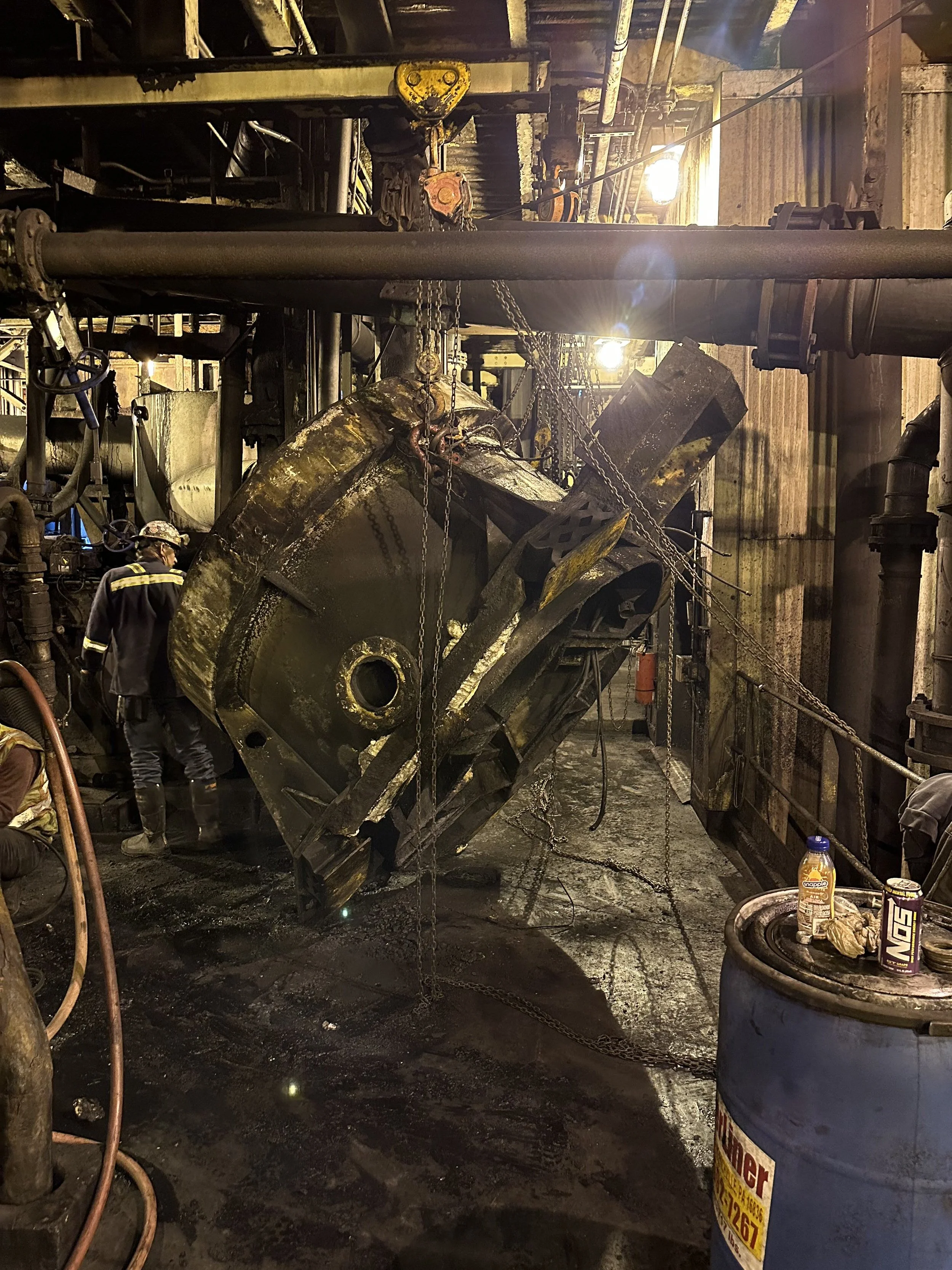 A worker in protective gear inspects large industrial machinery inside a factory or processing plant, with chains hanging around the machine, and a barrel in the foreground with a bottle and snack in it.