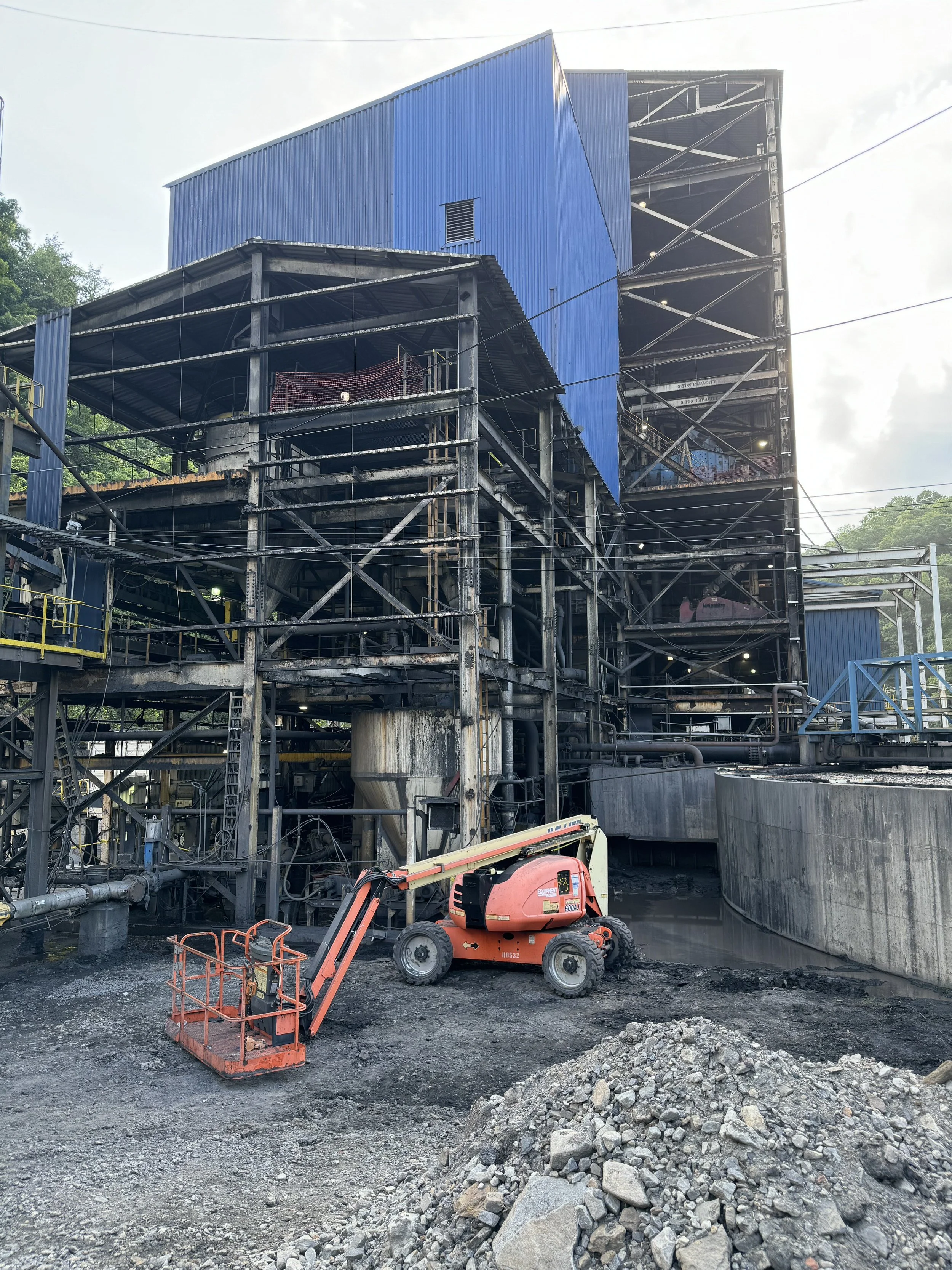 Construction site with a large industrial building under construction, featuring scaffolding and blue metal siding, a small orange lift equipment, and a pile of rocks and dirt in the foreground.