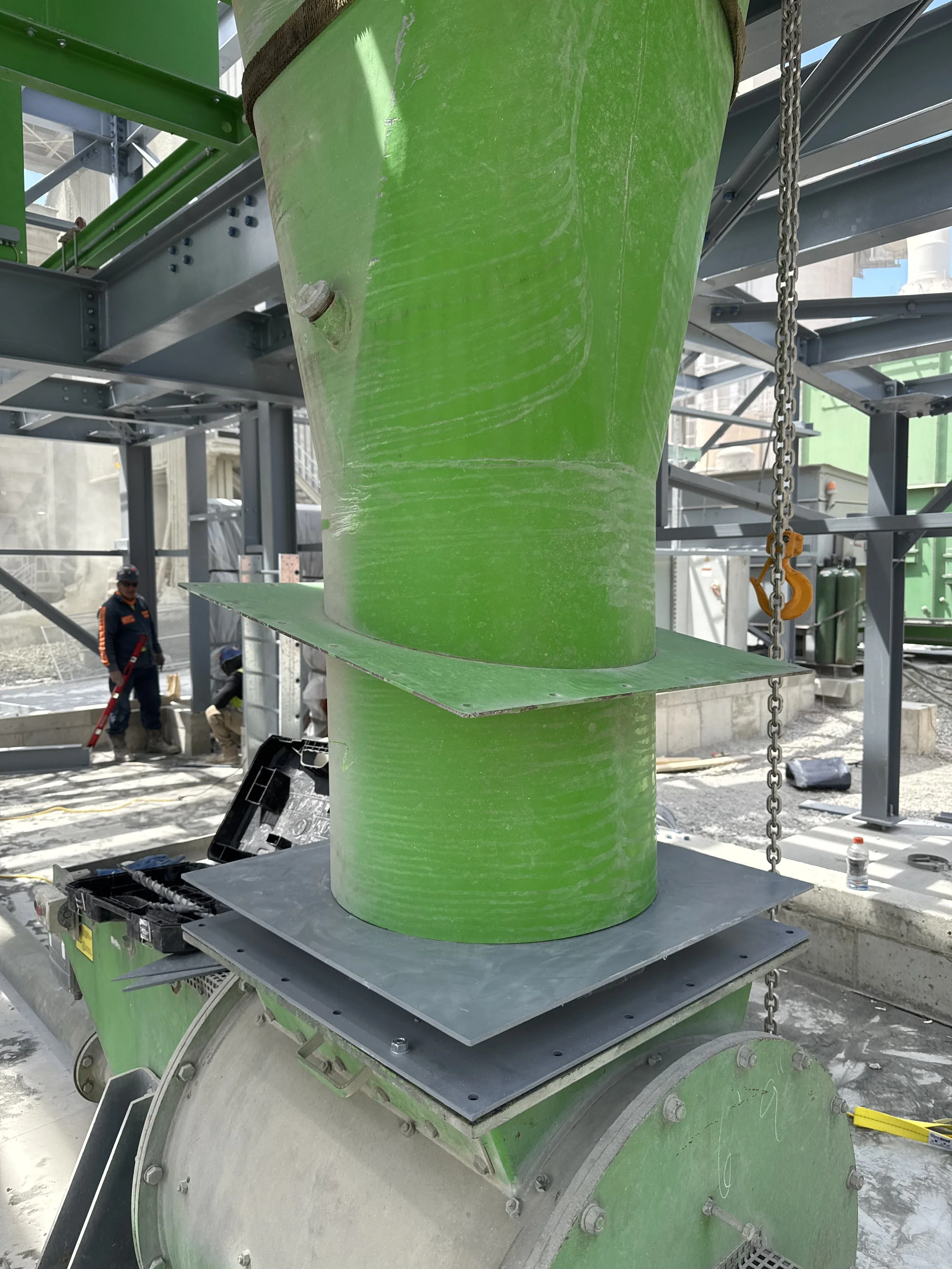Close-up view of a green industrial pipe at a construction site with workers and building structure in the background.