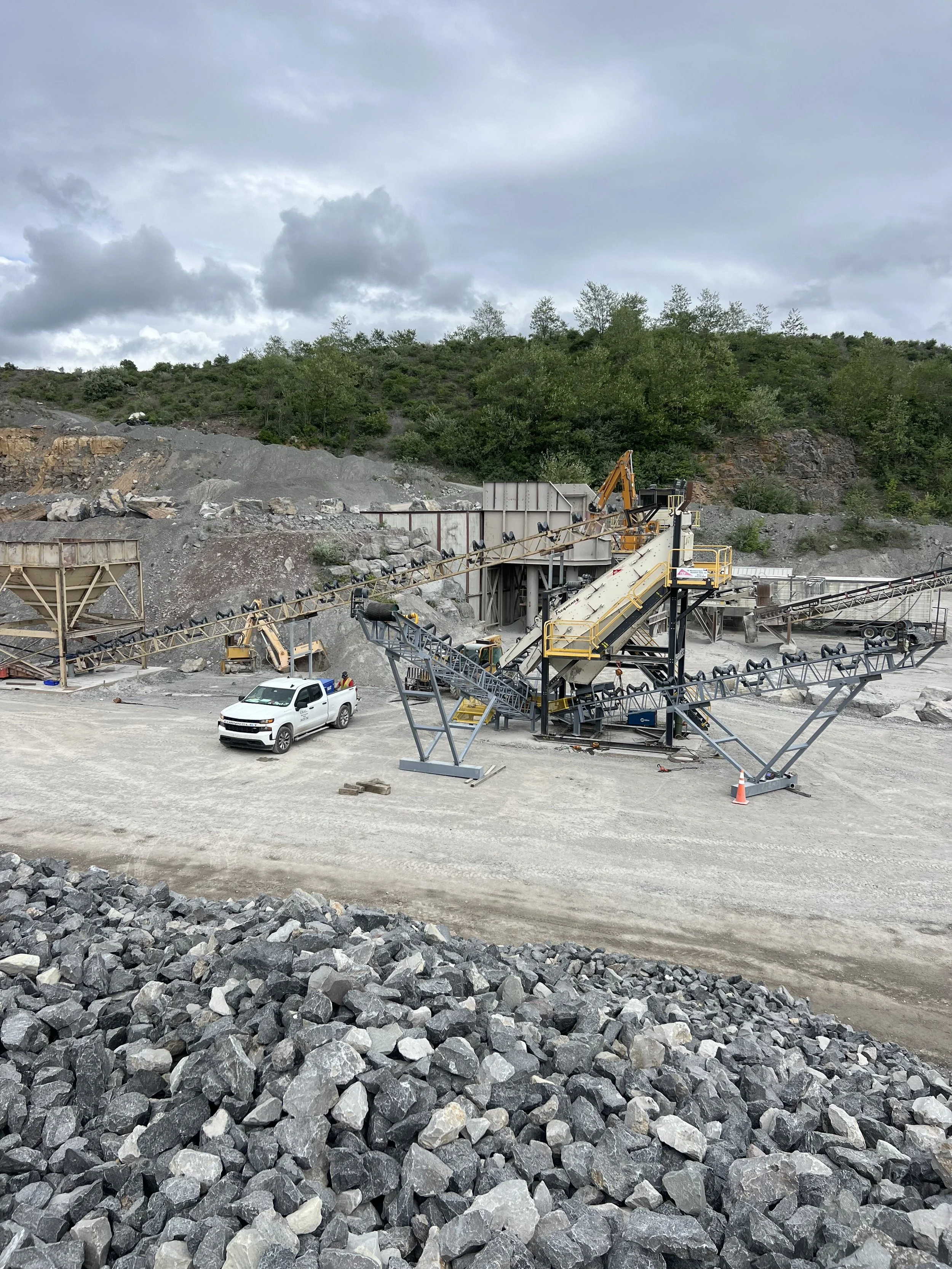 Rock quarry with conveyor belts, machinery, a white pickup truck, and a mountainous backdrop under cloudy sky.