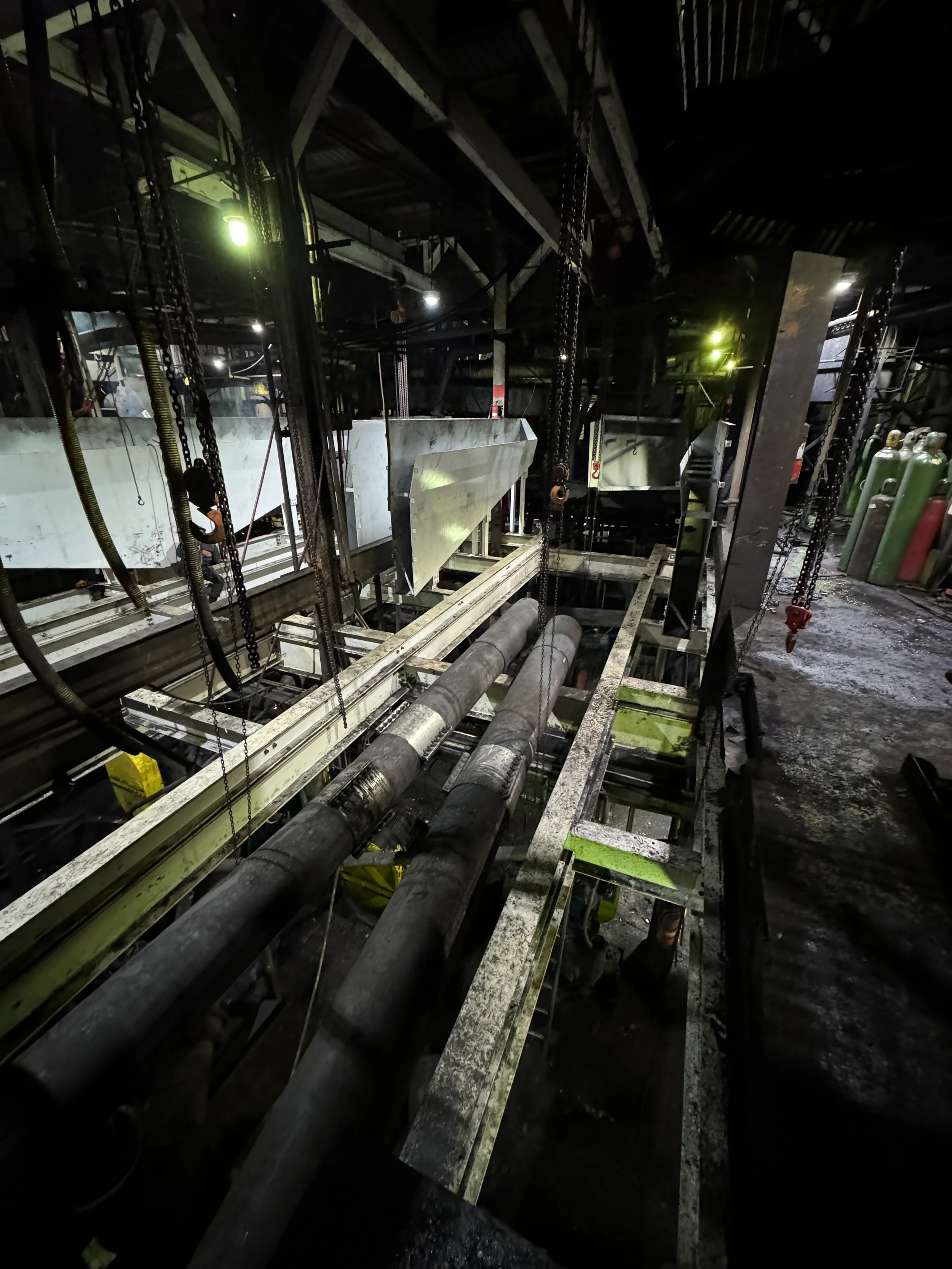 Dark industrial or construction site with pipes, chains, and metal framework, illuminated by faint green and white lights.