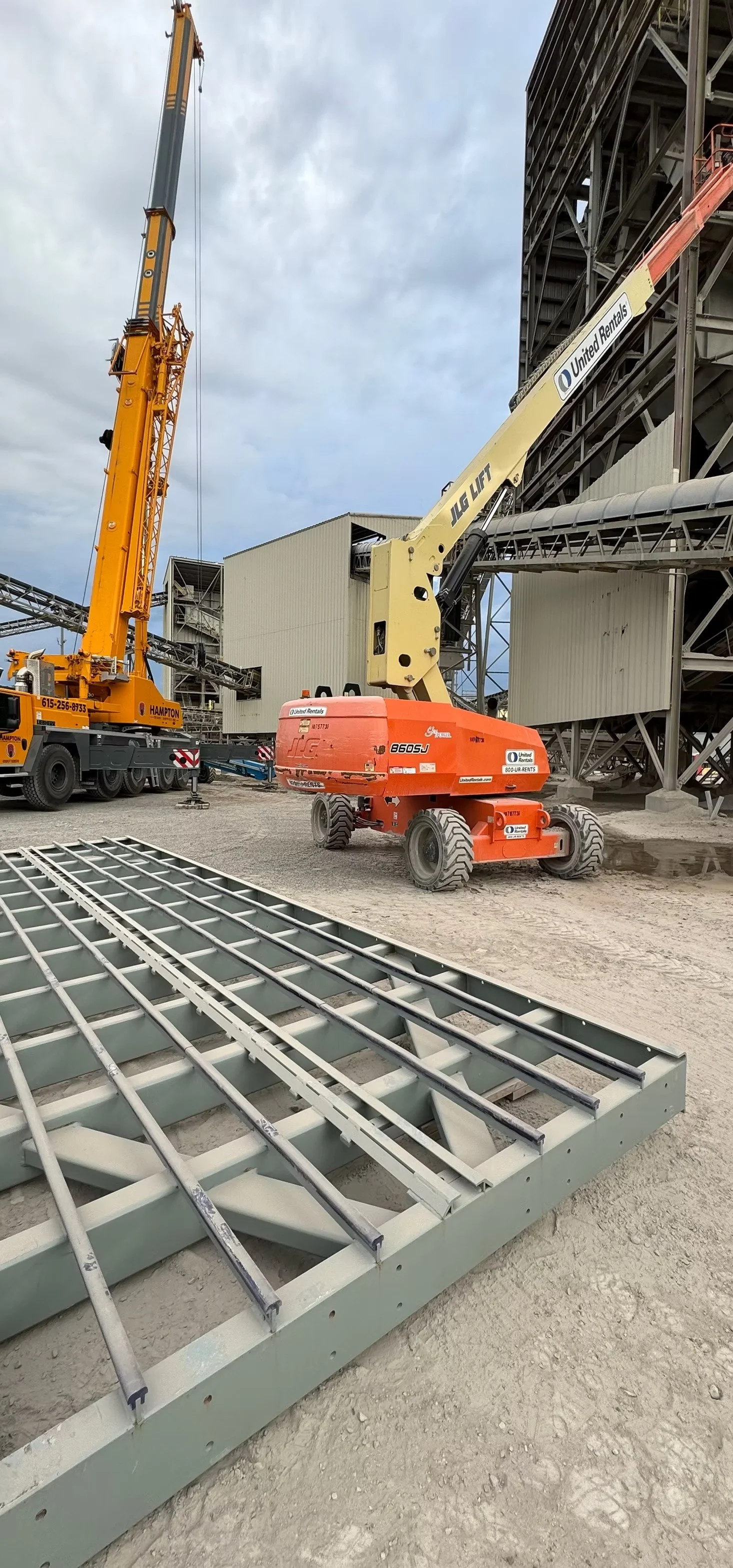 Construction site with a large orange JLG lift, a yellow crane, completed steel framework, and ground-level metal grid structures under cloudy sky.