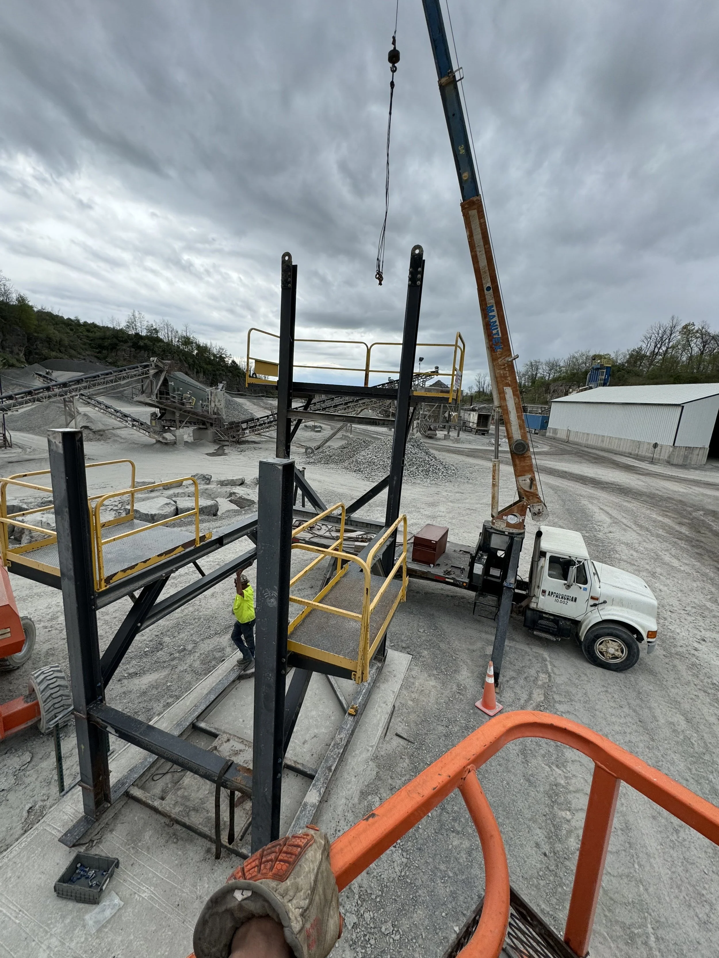 Construction site with a crane, work platform, and equipment, under cloudy sky, with workers and a gravel landscape.
