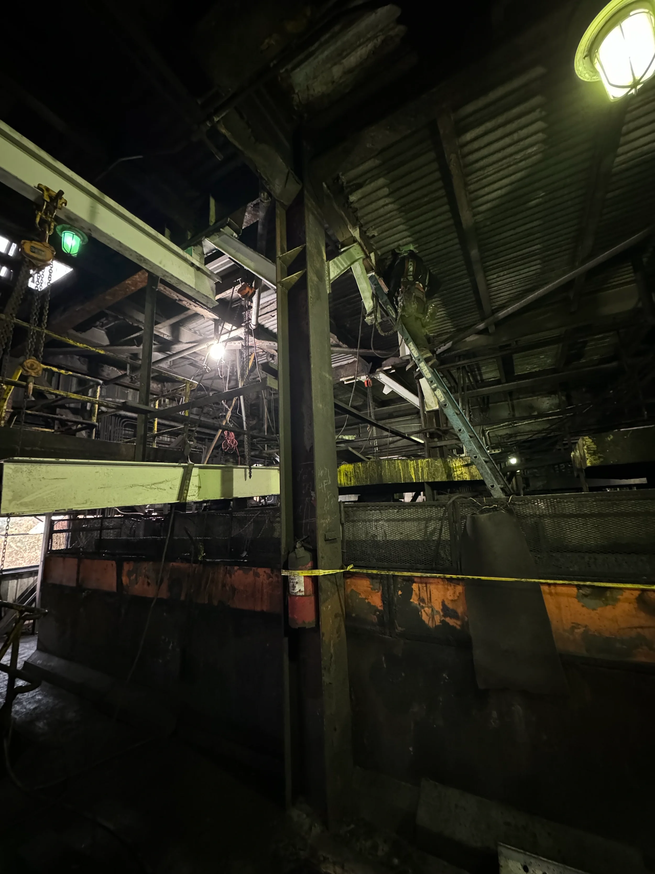 Dark indoor construction site with scaffolding, pipes, and industrial equipment, illuminated by hanging lights and a small green safety light. A worker in safety gear is visible in the background.