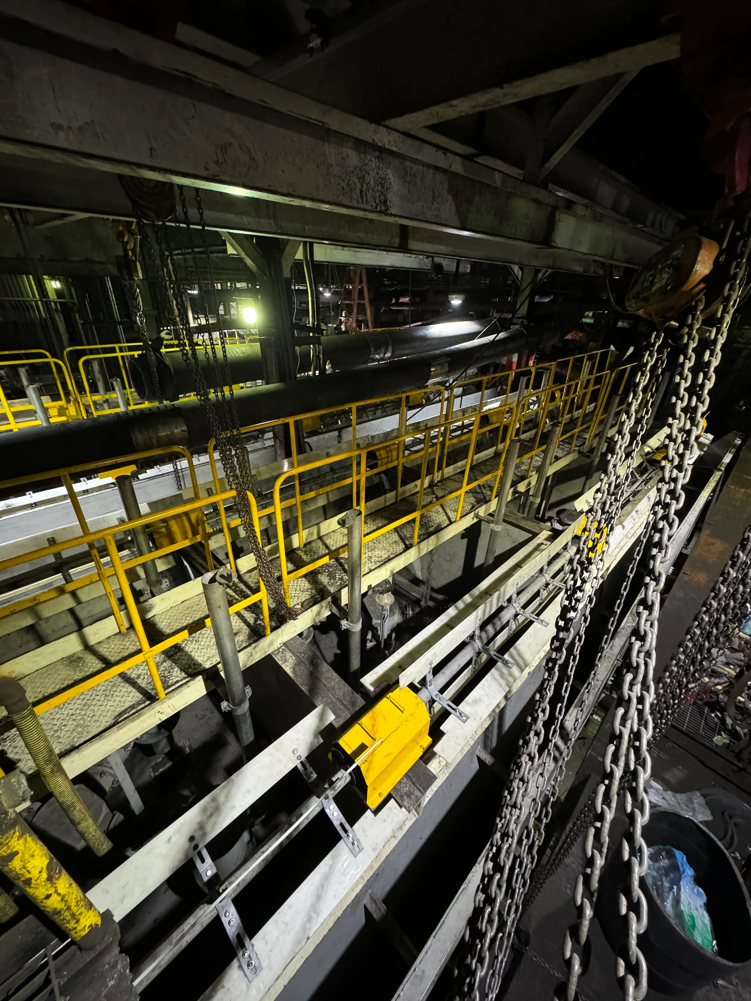 Industrial scene featuring pipes, yellow safety railings, chains, and metal beams in a dark setting, possibly a factory or power plant.