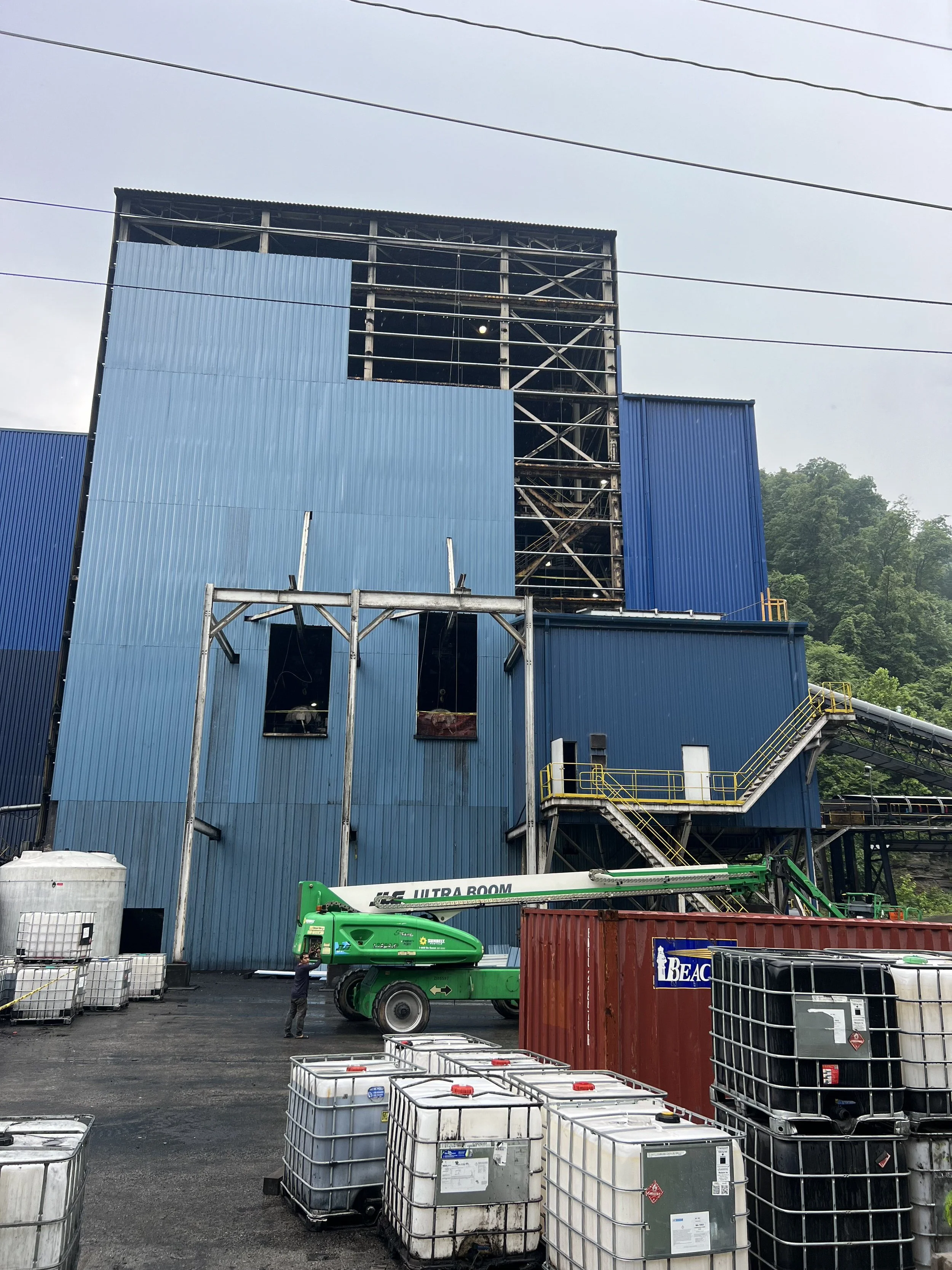 Construction site with a blue building structure under construction, multiple white and black storage tanks, a green construction lift, and a person standing nearby, with trees in the background.