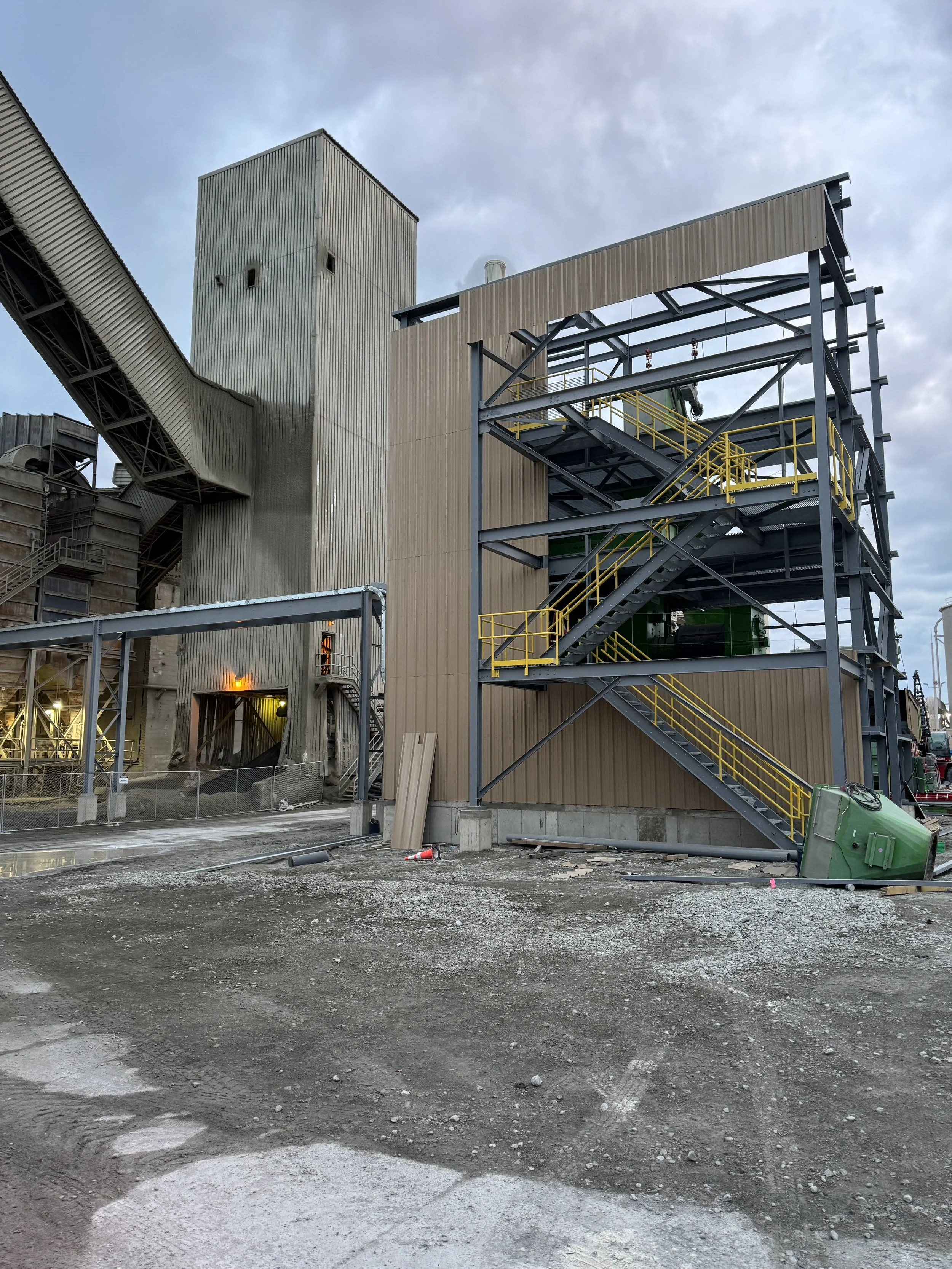 Construction site with industrial buildings featuring metal structures, stairs with yellow railings, and a cloudy sky.