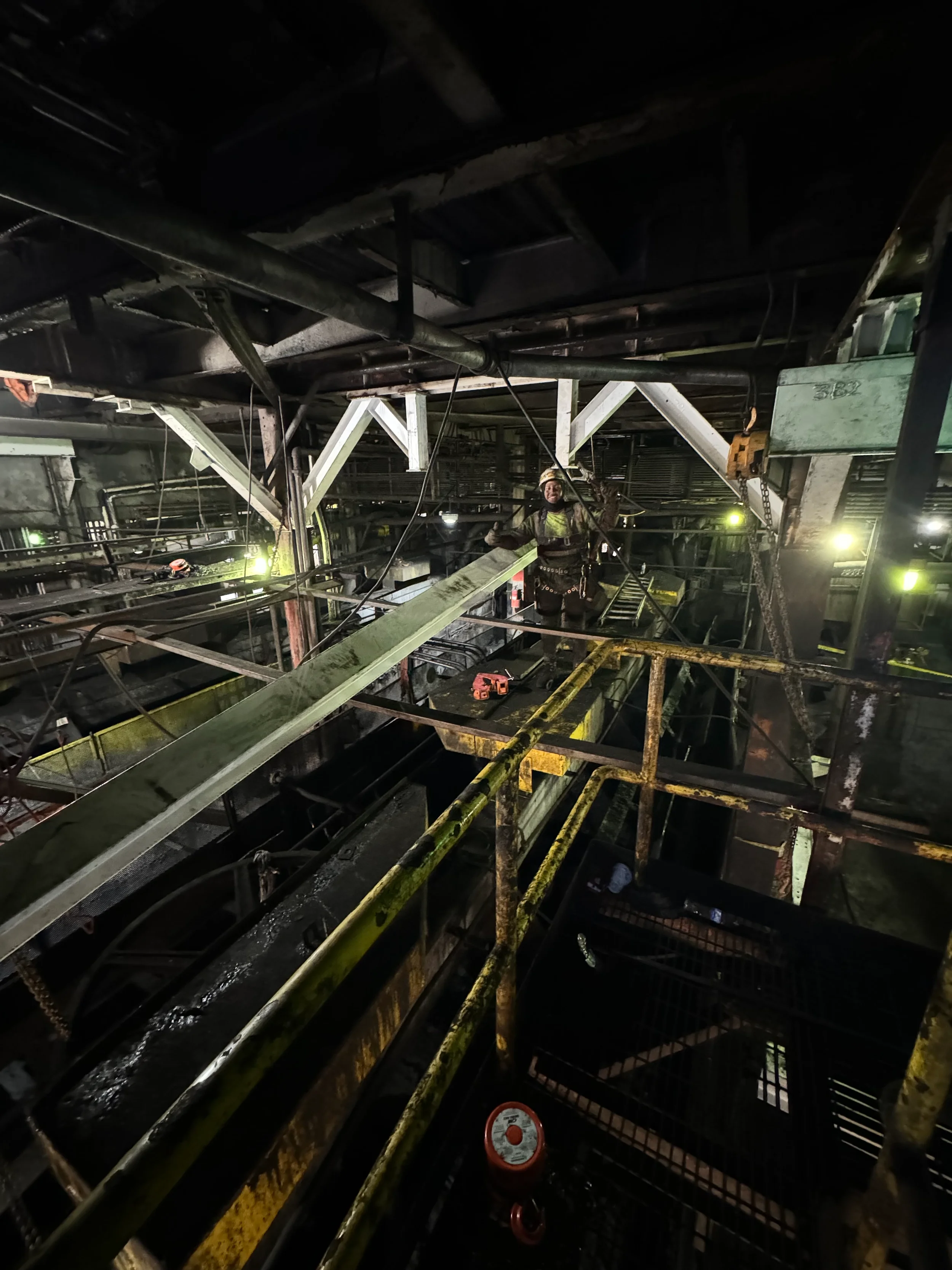Worker in industrial setting on scaffolding at night, wearing safety harness and helmet, surrounded by metal pipes and machinery.