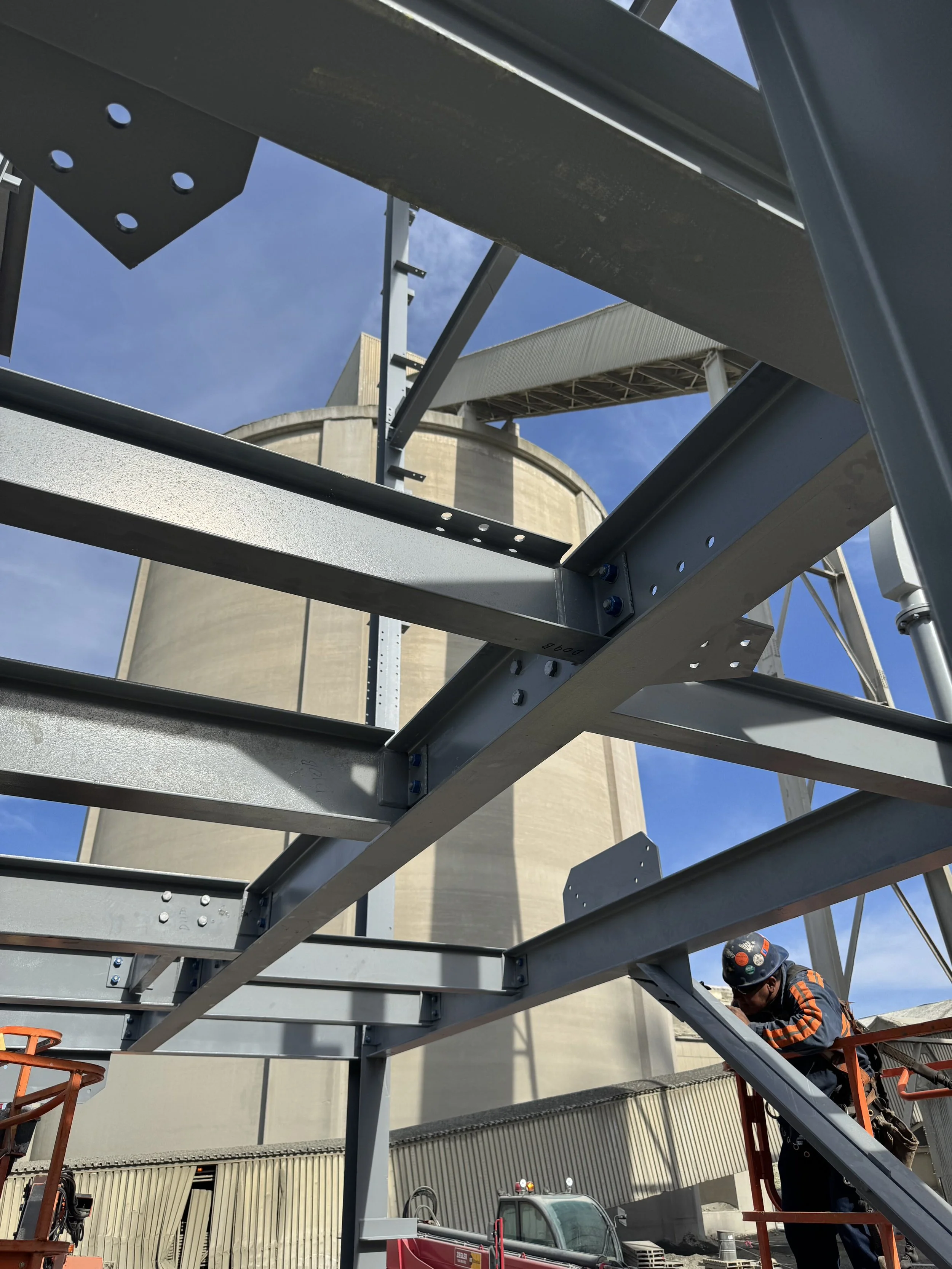 Construction worker wearing safety gear inspecting a steel framework structure at a construction site with a large building and blue sky in the background.