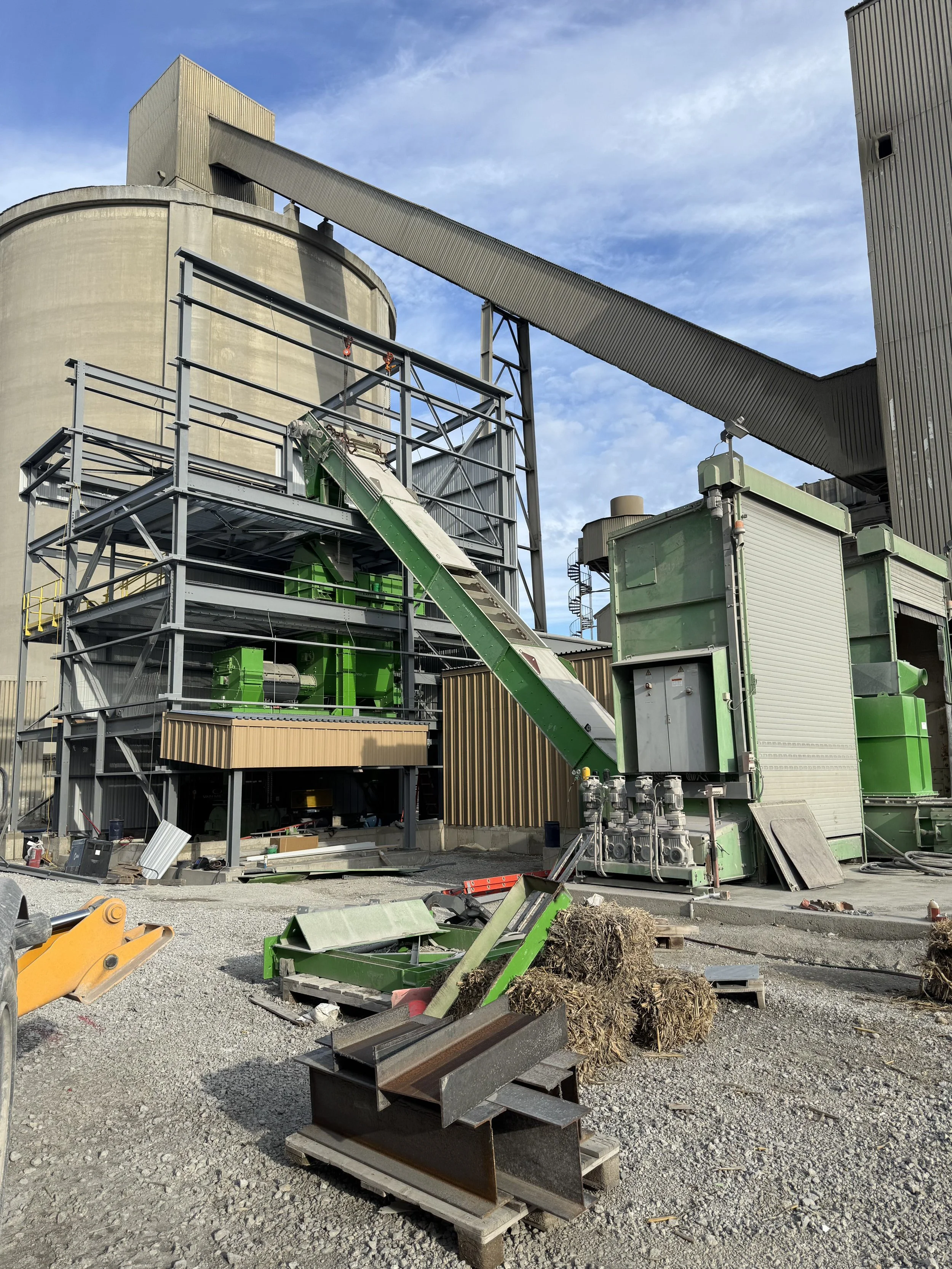 Industrial construction site with machinery, metal structures, and building elements under a partly cloudy sky.