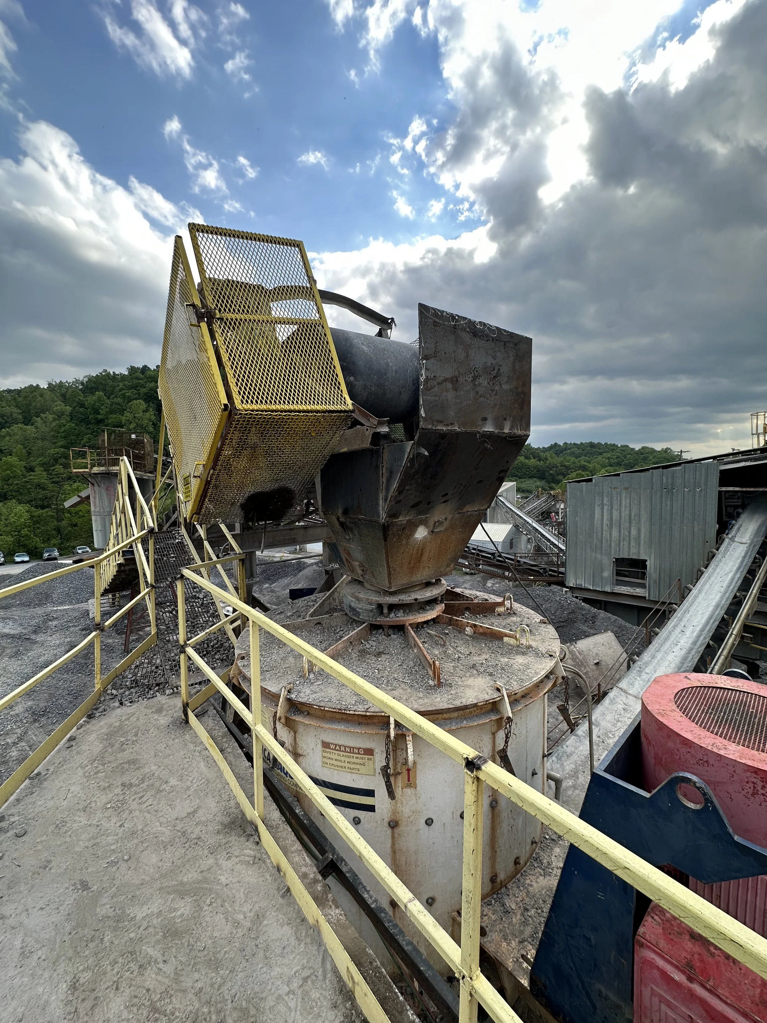 Large industrial machine outdoors under a partly cloudy sky, surrounded by a yellow safety railing, with a forested hillside in the background.
