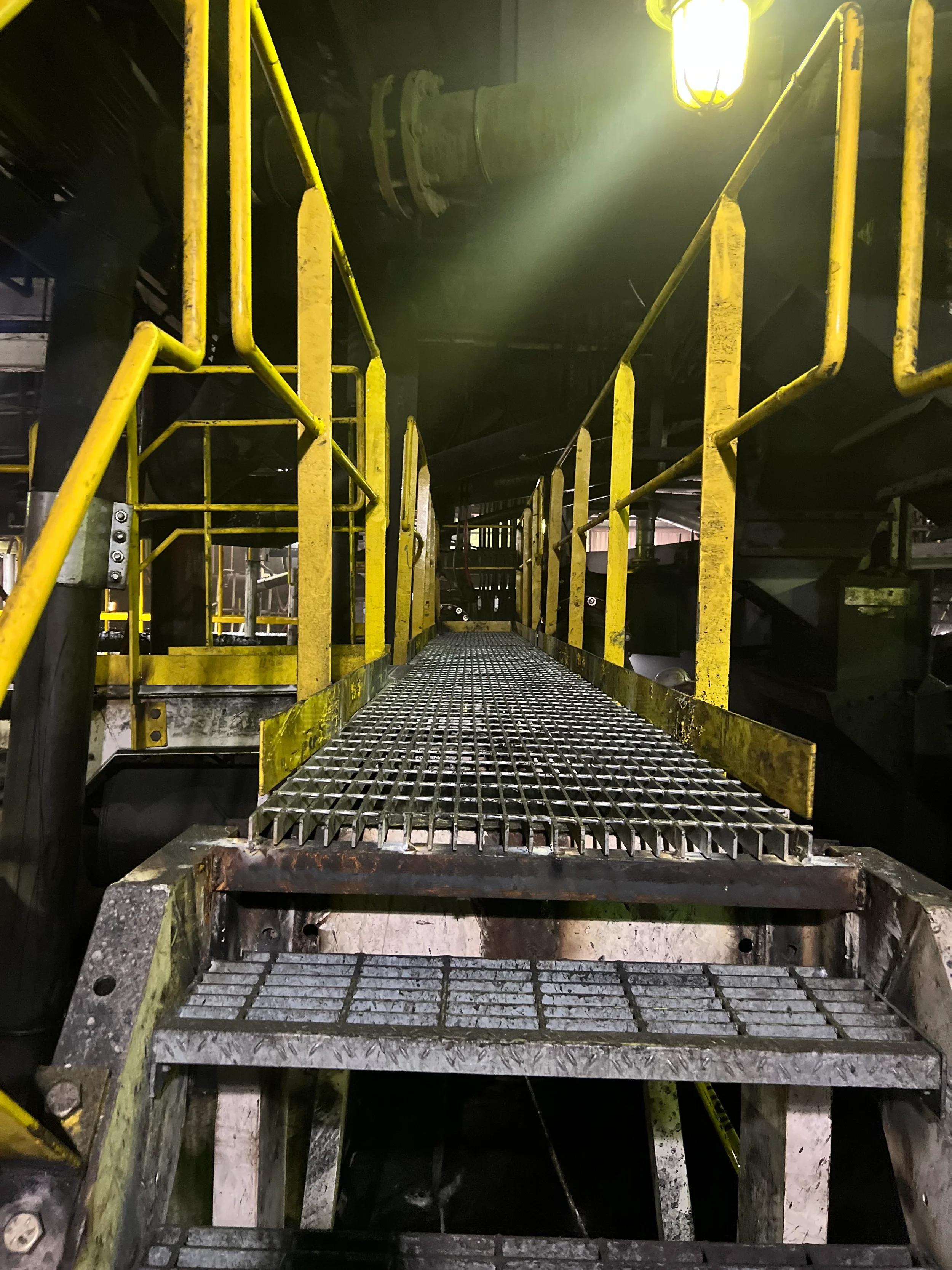 Industrial metal staircase with yellow railing, leading down into a dark, industrial space, illuminated by a bright light at the top right corner.