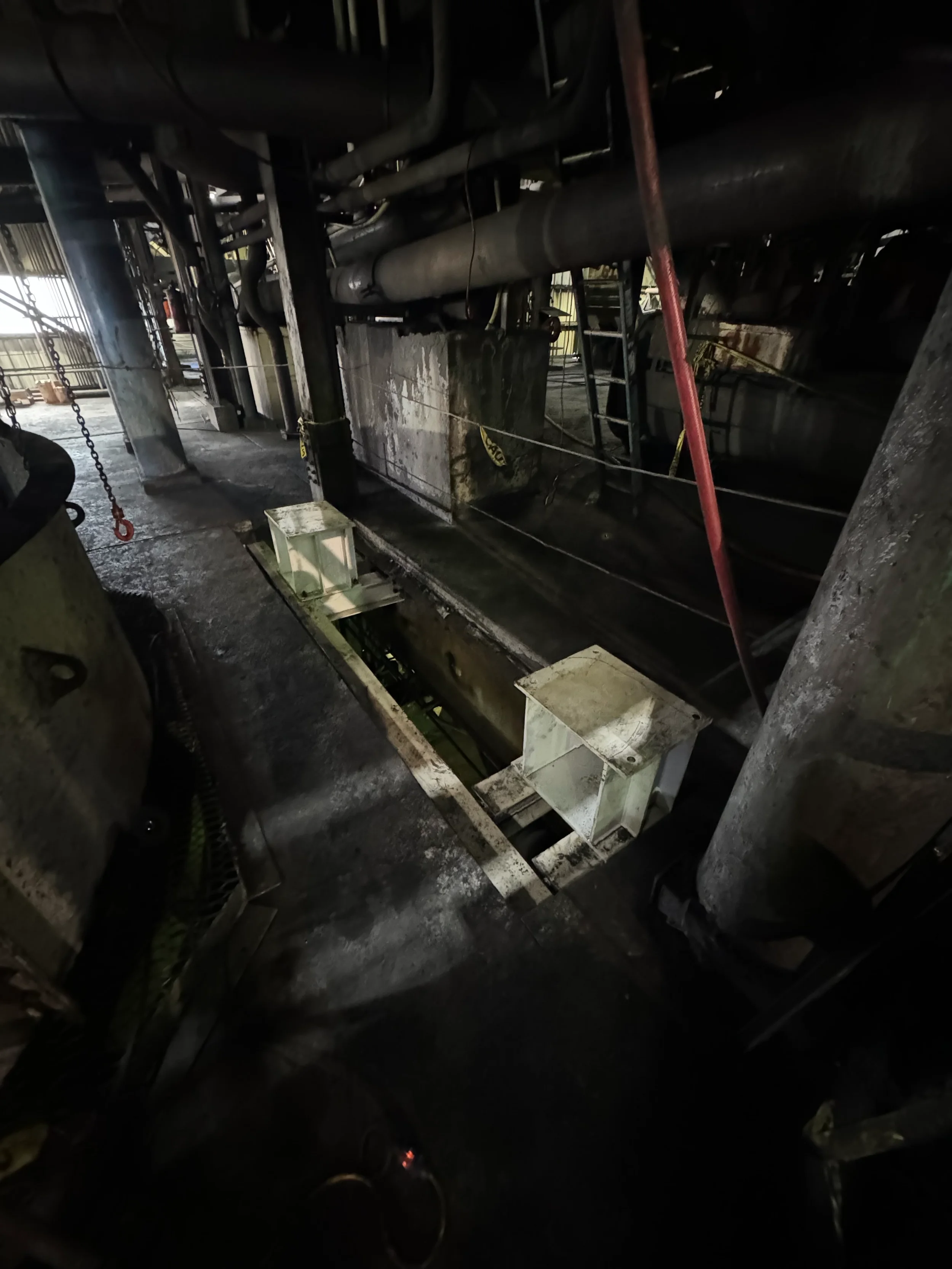 Dark industrial interior with pipes, metal structures, and a ladder, illuminated by dim light.