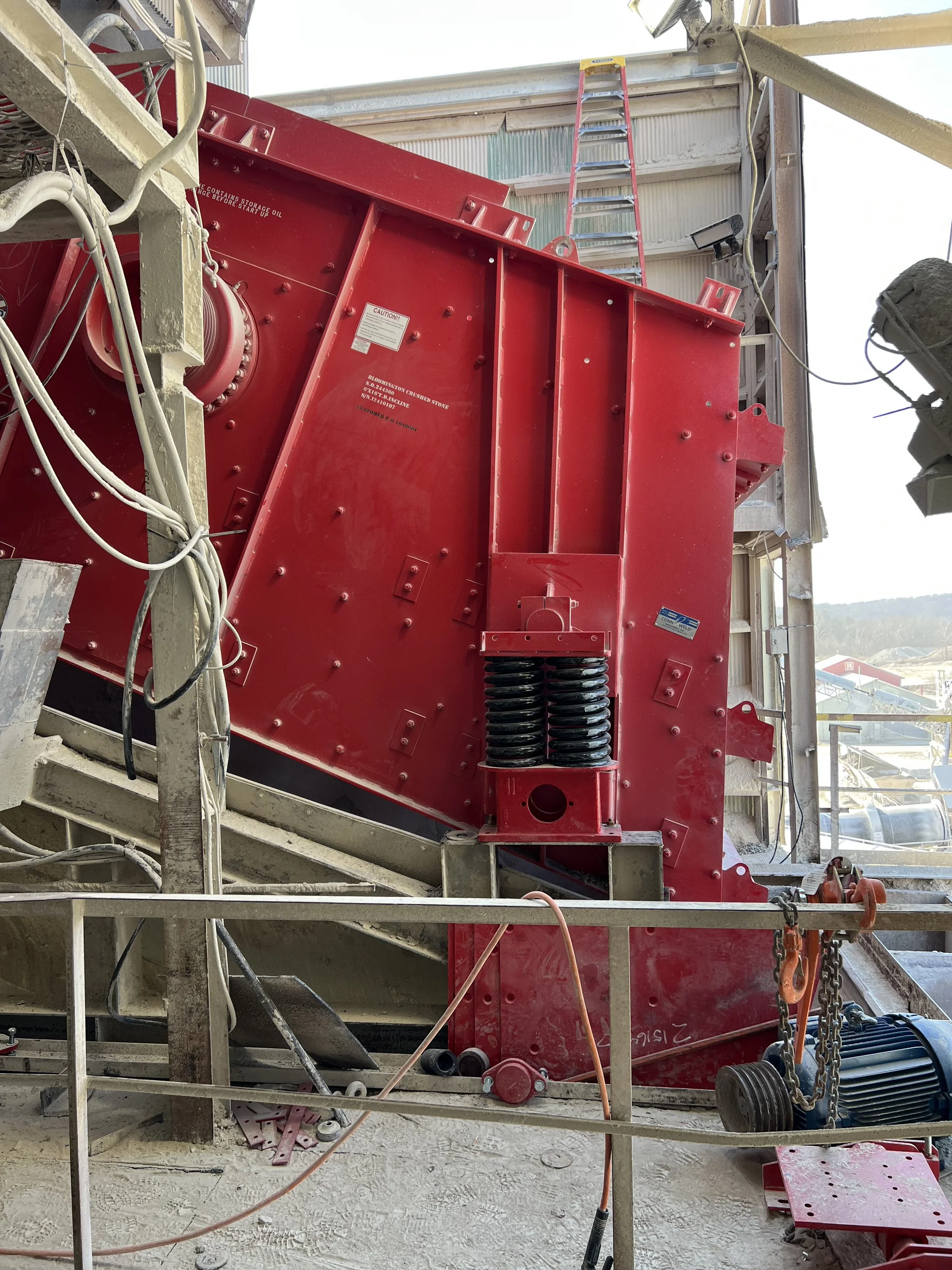 Close-up of construction equipment including a large red machine, wiring, a ladder, and other construction materials at a building site with an open wall and distant view of a landscape and buildings.