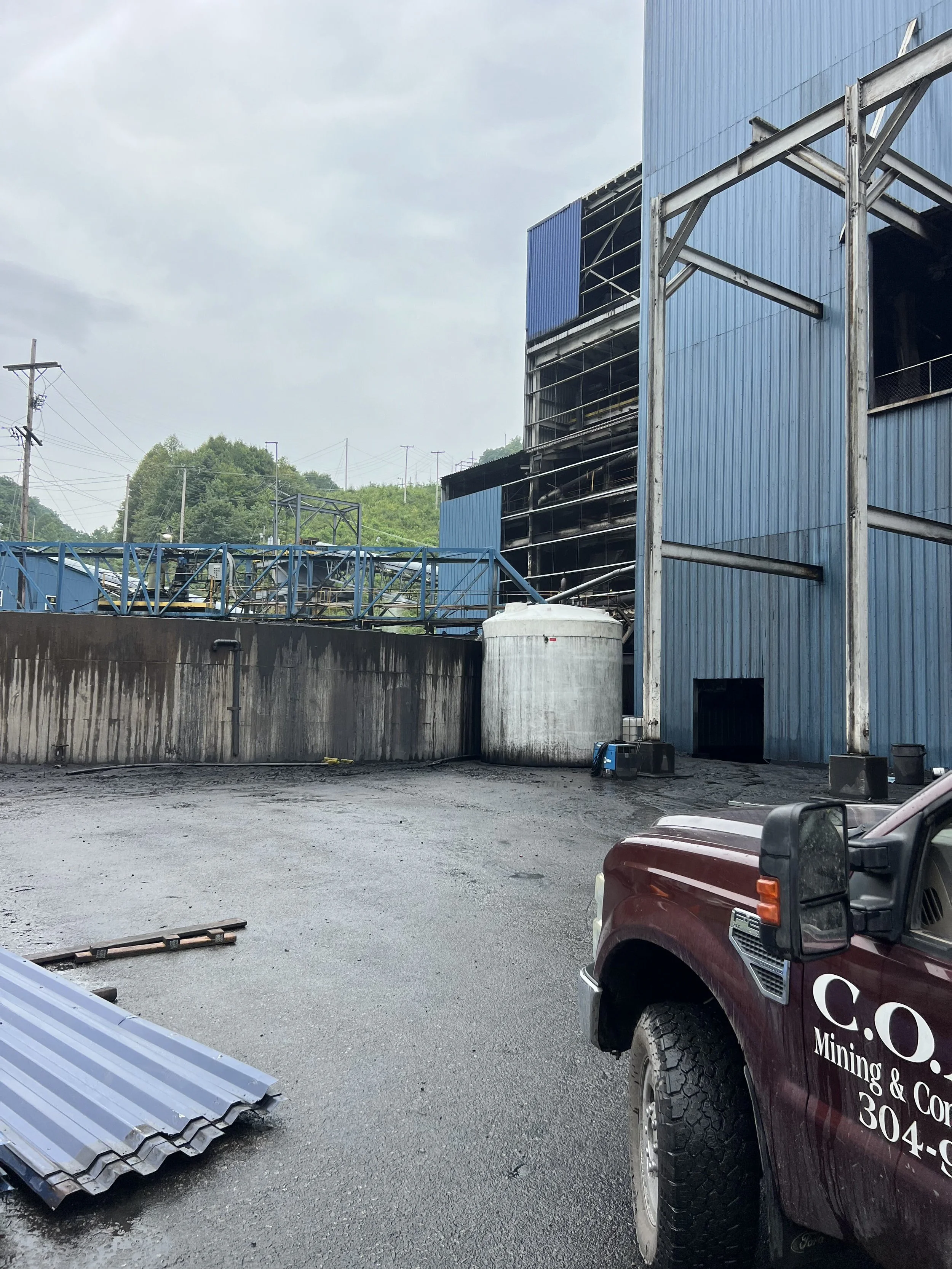 View of an industrial area with a blue metal building, a gray cylindrical tank, a maroon company pickup truck, a few metal sheets, and utility poles with wires. Overcast sky in the background.