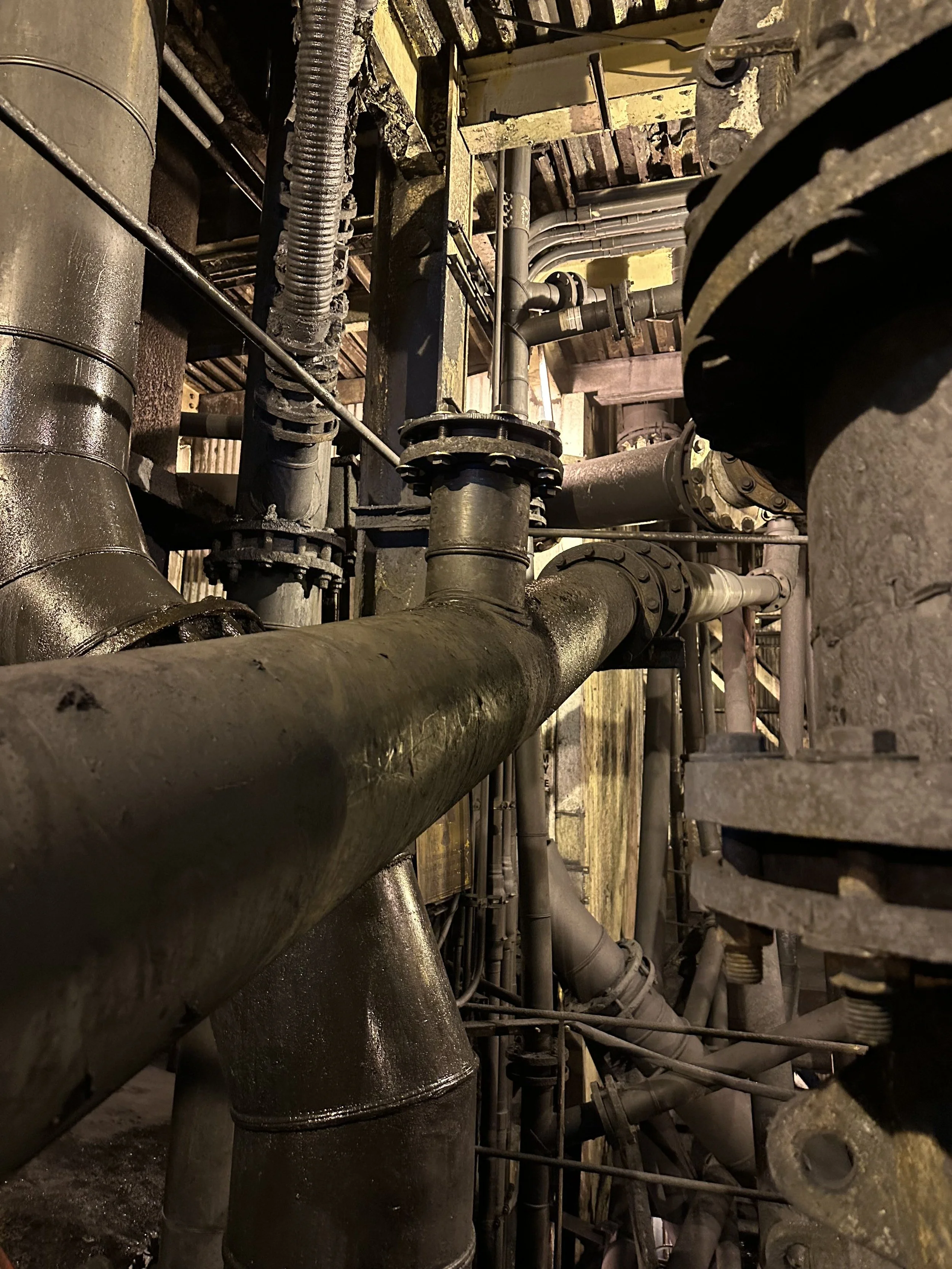 A complex network of industrial pipes, valves, and fittings, some of which are connected to a large, rounded metal tank or vessel, in a dimly lit industrial setting.