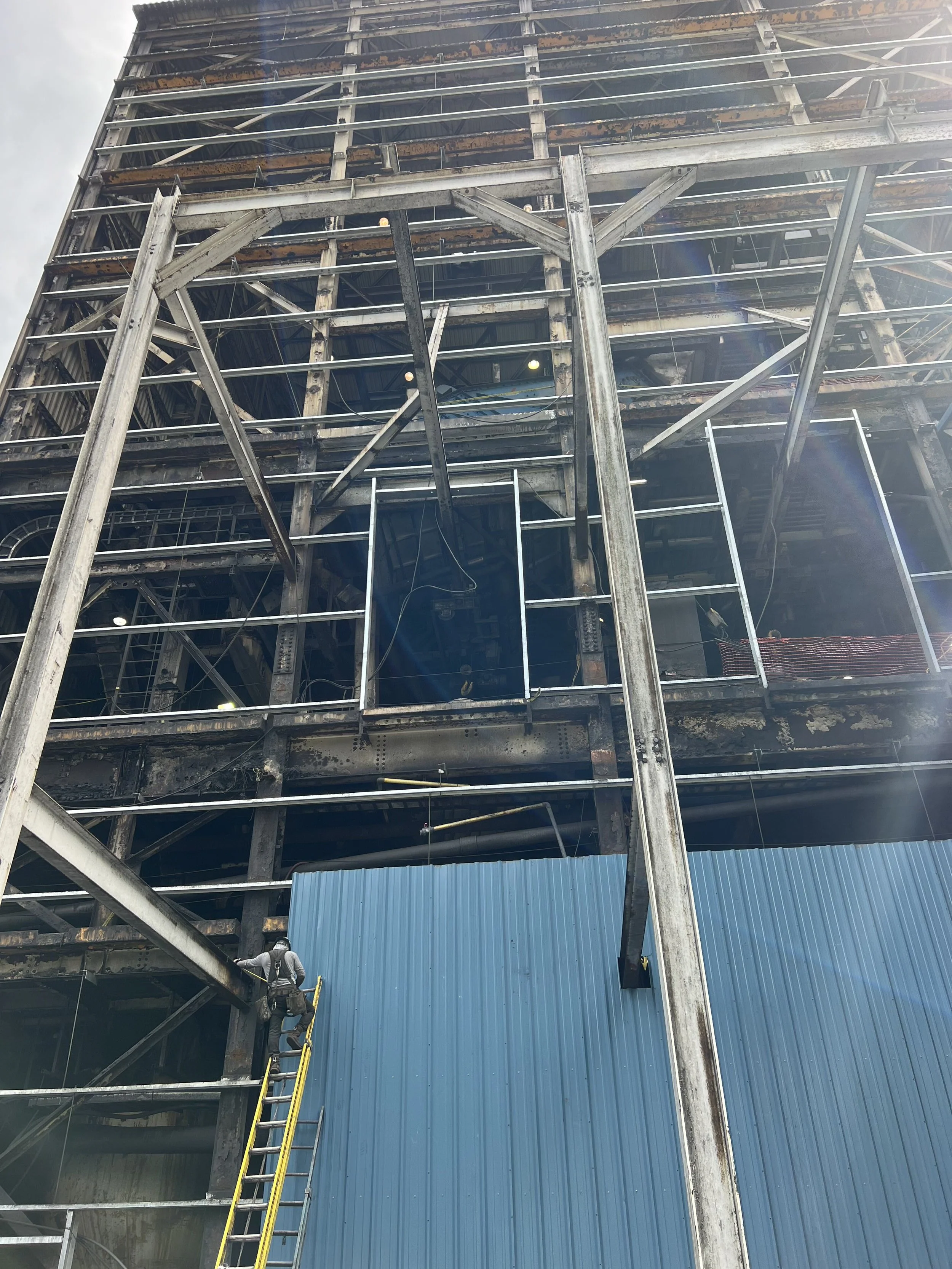 A multi-story building under construction with a steel frame structure. A worker is standing on a yellow ladder at the bottom left, wearing a helmet and harness. Some areas have blue metal sheeting, and there are adjacent construction materials and e