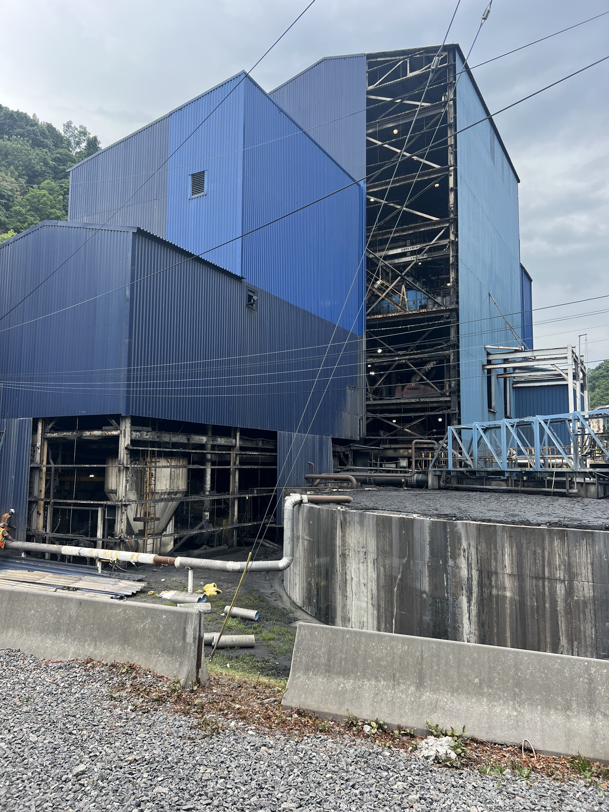 Large industrial building with blue metal siding; some sections are under construction with visible steel framework; concrete barriers and gravel in foreground; utility pipes and wires around the site; cloudy sky in the background.