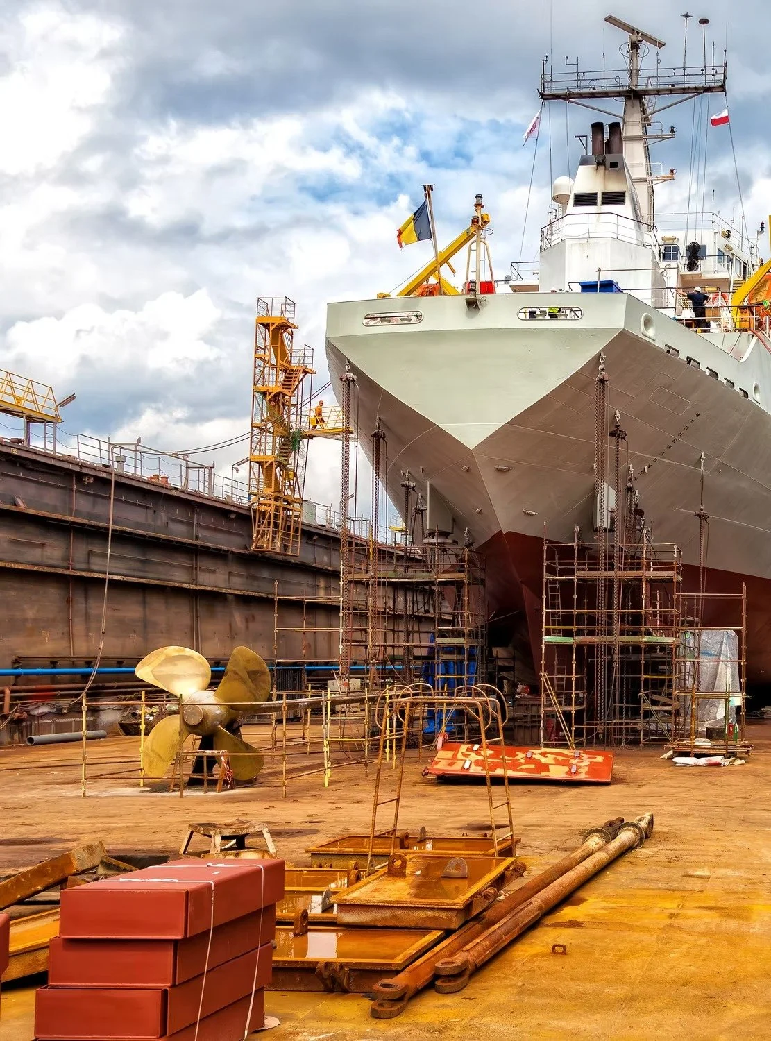 View of a ship in a shipyard with scaffolding, ladders, a boat propeller, and construction materials around.